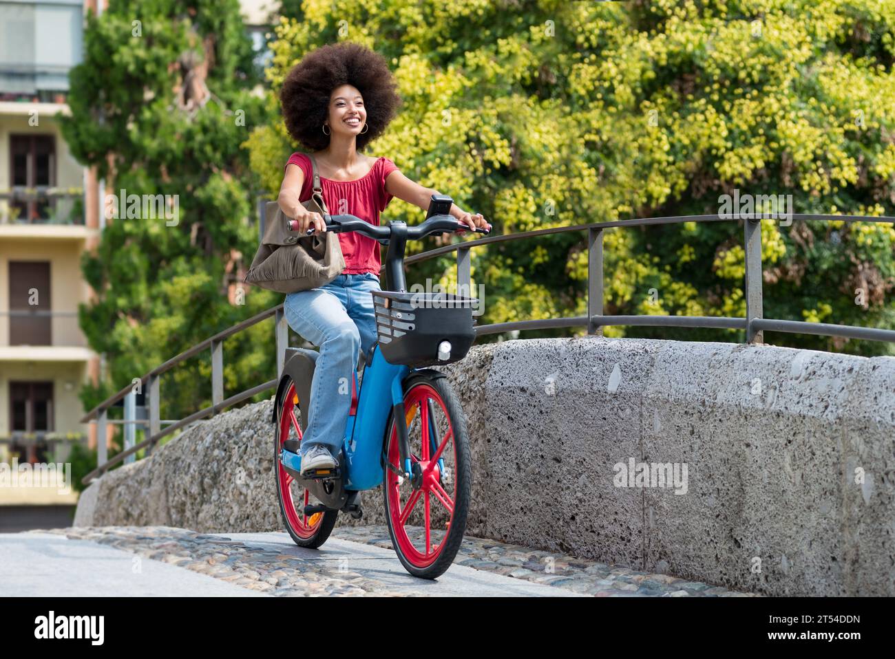 Vista laterale di una giovane donna marocchina sorridente con una borsa che guarda dall'altra mentre si guida in bicicletta elettrica sul ponte cittadino con ringhiere contro la t verde sfocata Foto Stock