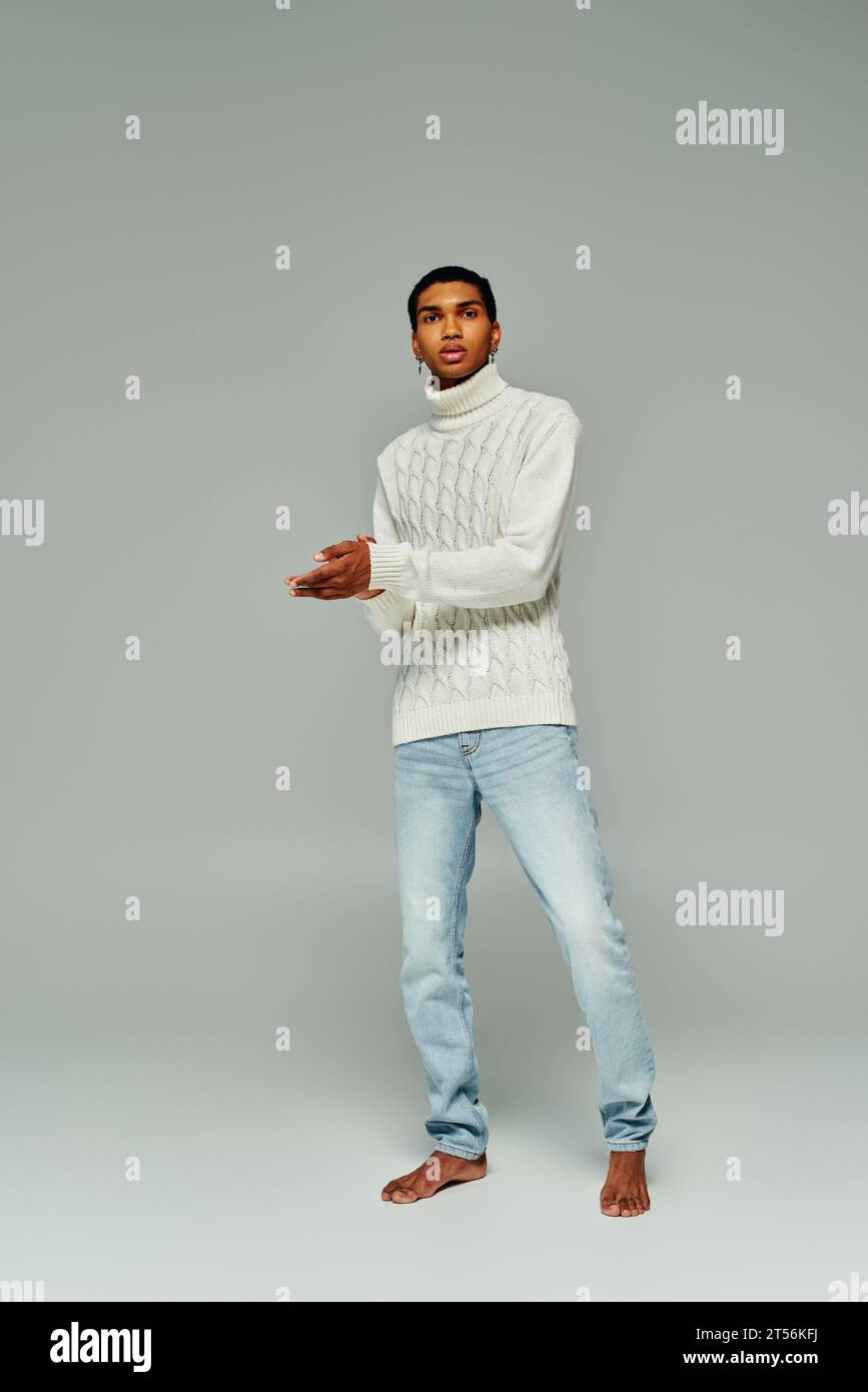 uomo afroamericano dall'aspetto gradevole in maglione bianco con accessori in posa su sfondo grigio, moda Foto Stock