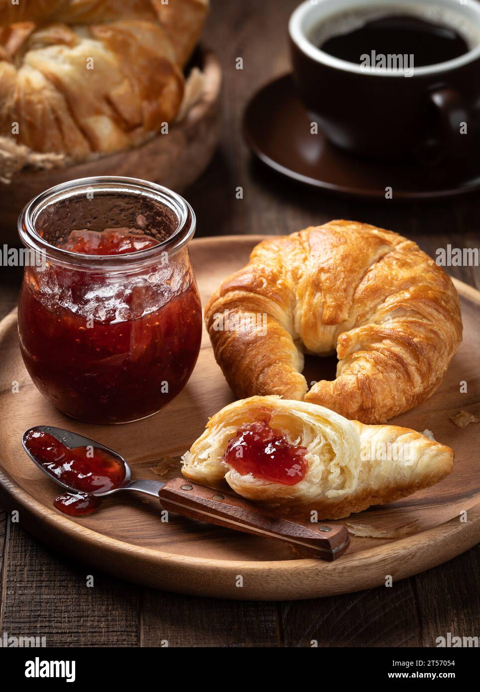 Croissant con confetture di fragole su piatto di legno con croissant e tazza di caffè sullo sfondo Foto Stock