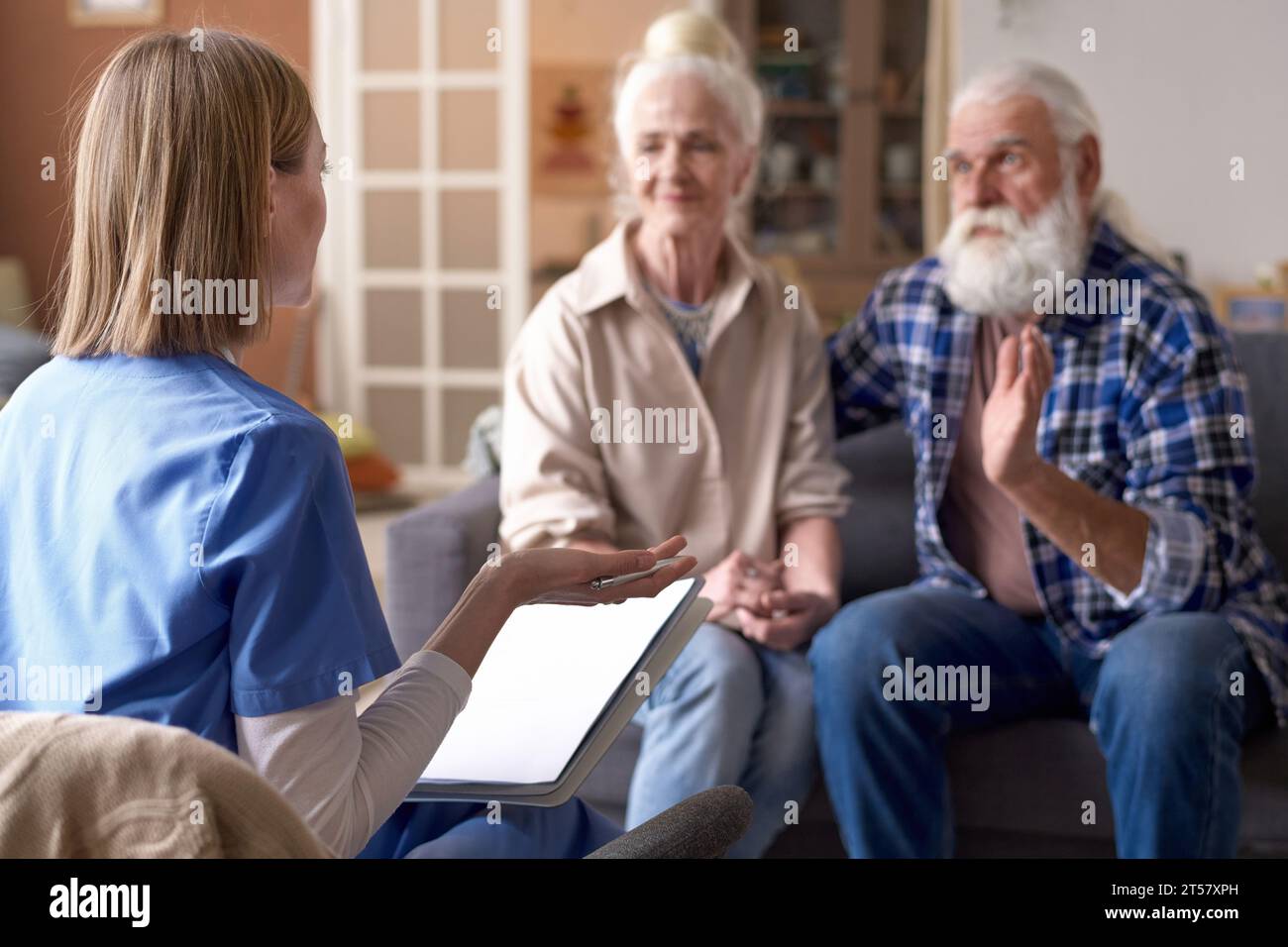 Vista posteriore di un giovane assistente sociale che parla con una coppia anziana nel salotto di casa Foto Stock