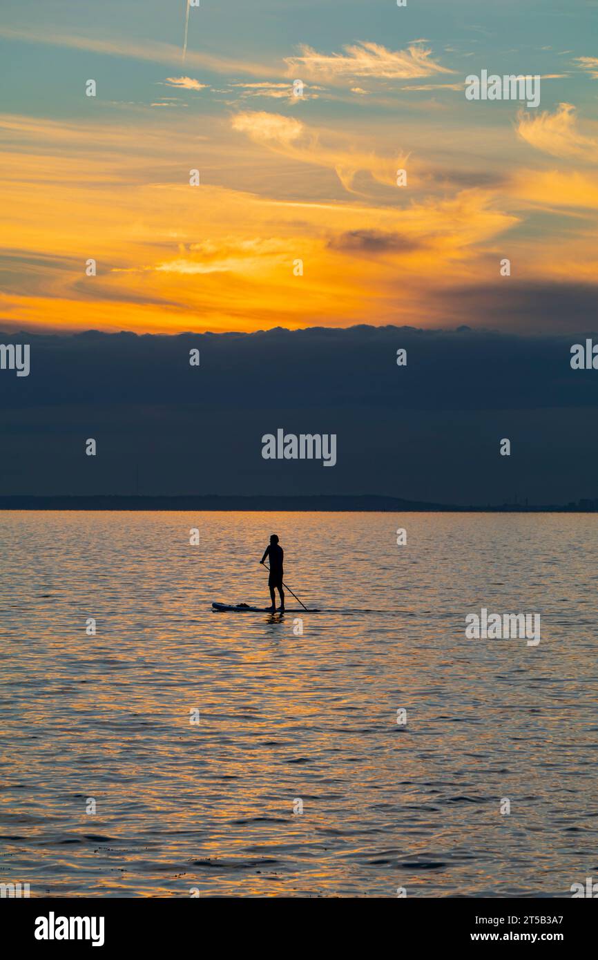 Il sole che tramonta illumina la nuvola all'orizzonte mentre un paddle boarder passa lentamente Foto Stock