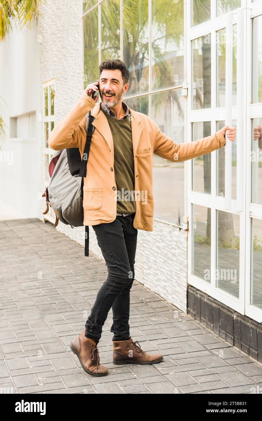 Uomo moderno sorridente con lo zaino che parla con il cellulare mentre apre la porta di vetro Foto Stock