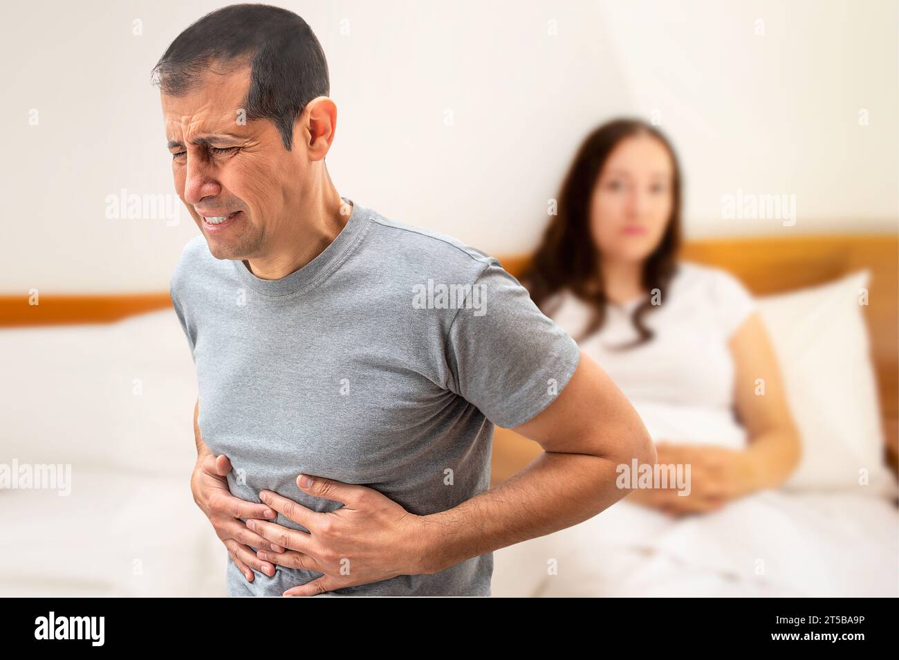 Un uomo che soffre di crampi allo stomaco seduto sul suo letto vicino a sua moglie Foto Stock
