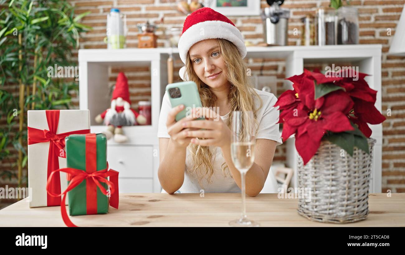 Giovane bionda che festeggia il natale con uno smartphone e beve champagne in sala da pranzo Foto Stock