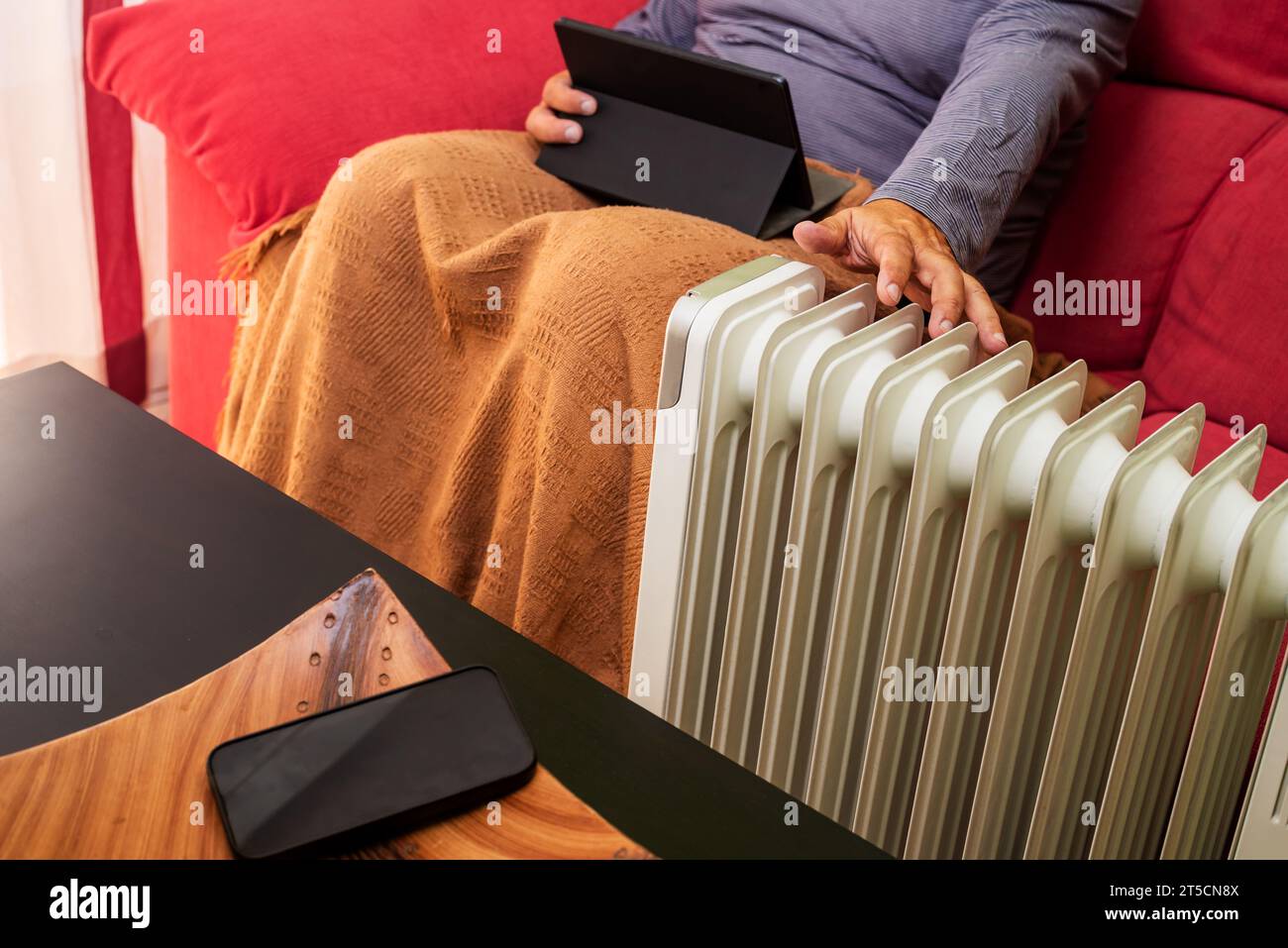 Uomo in pigiama sul divano a casa e coperto da una piccola coperta con un tablet in una mano e l'altra posta davanti al radiatore per riscaldarsi Foto Stock