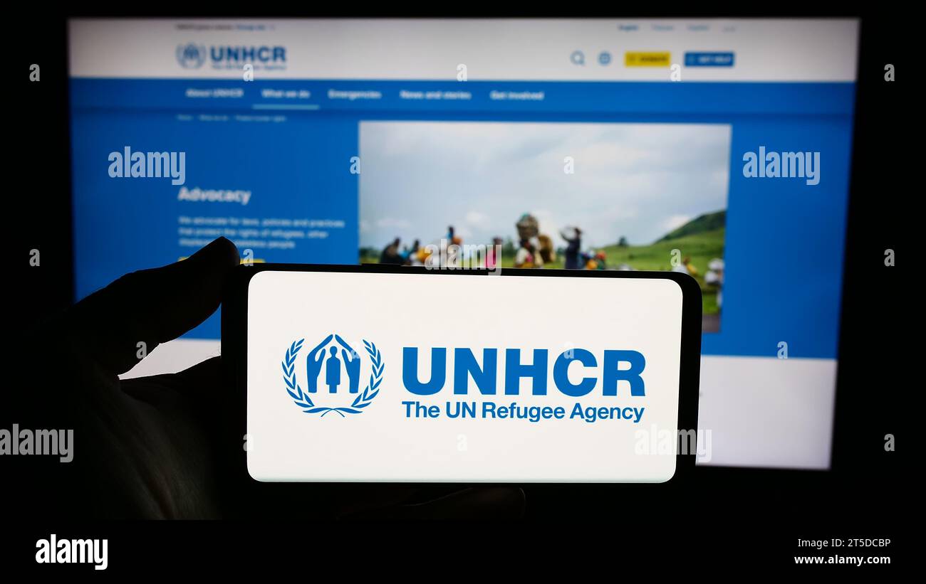 Persona che possiede uno smartphone con il logo dell'alto Commissariato delle Nazioni Unite per i rifugiati (UNHCR) davanti al sito web. Concentrarsi sul display del telefono. Foto Stock