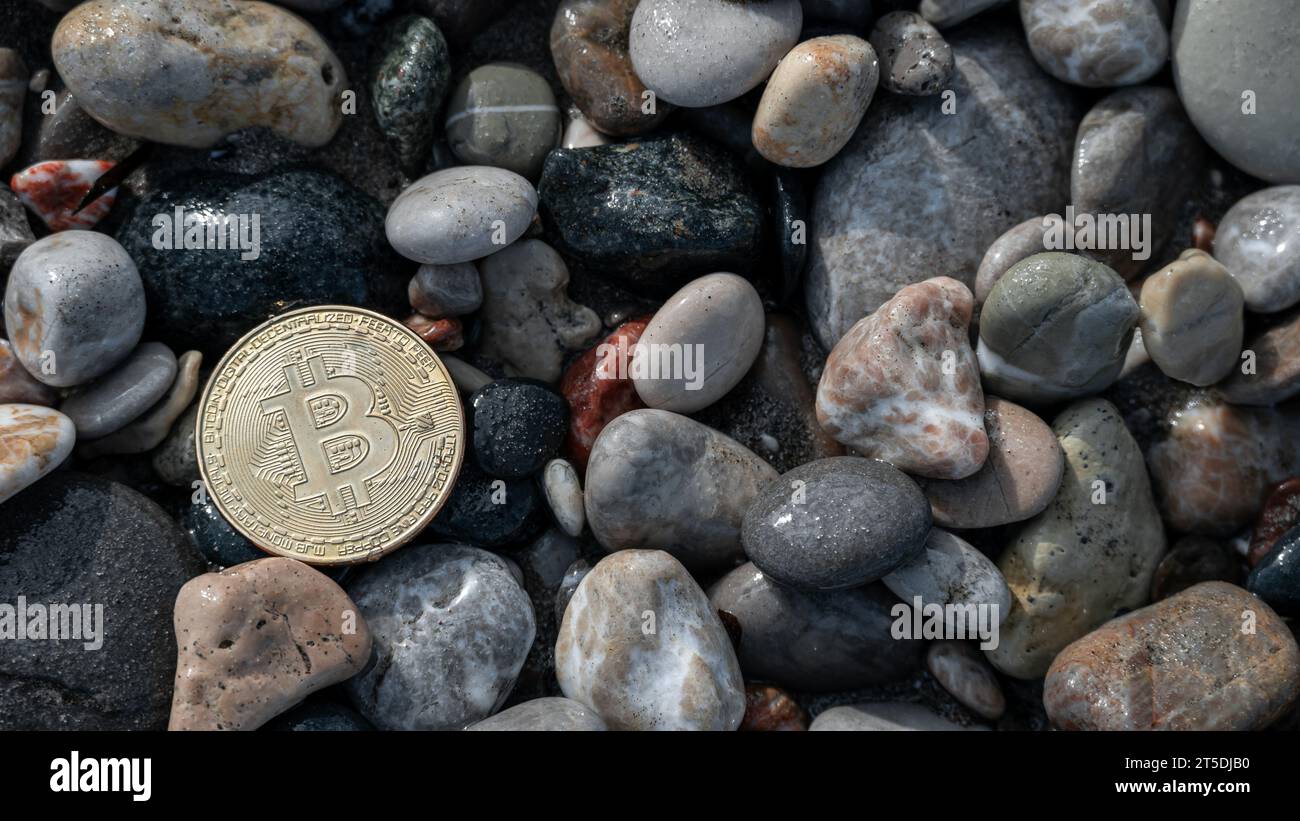 Moneta Bitcoin dorata posta su uno sfondo di grandi pietre grigie lungo la costa, a simboleggiare la stabilità contemporanea della moneta elettronica. Foto Stock
