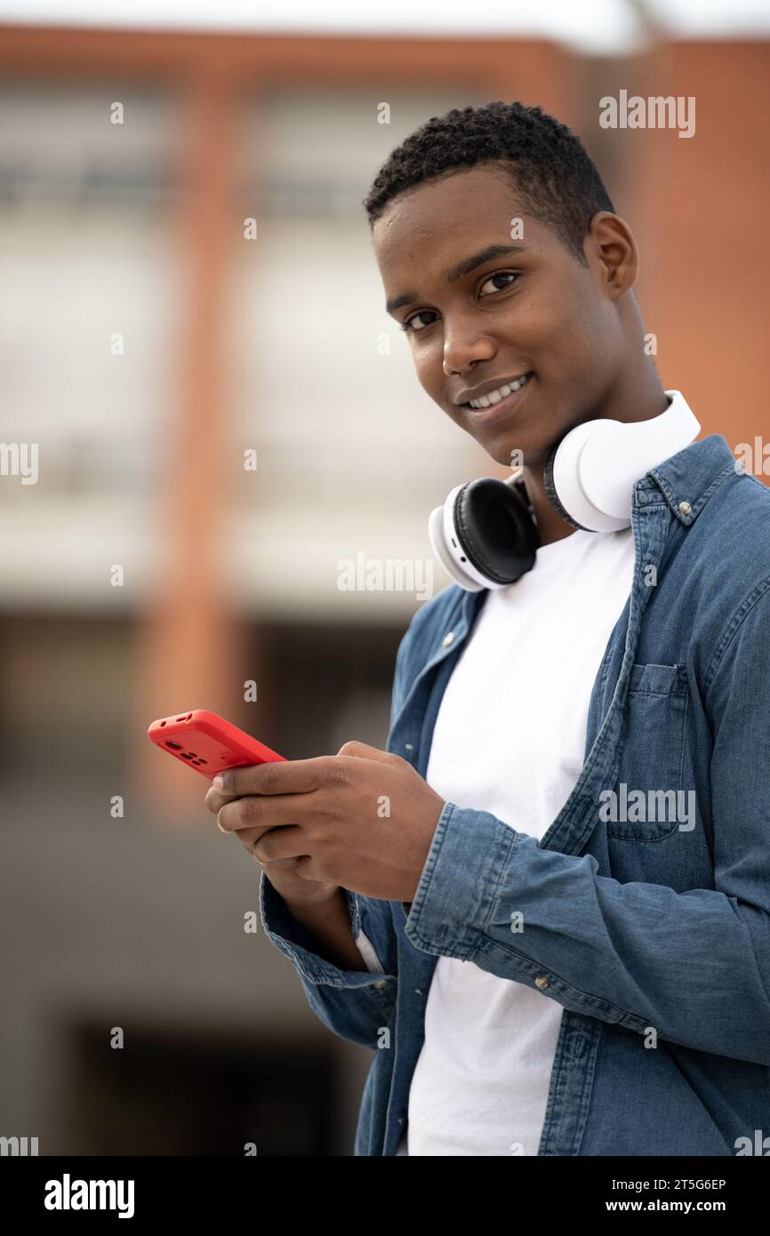 Un giovane ragazzo sorridente che guarda la fotocamera si rilassa usando il telefono.un uomo millenario che sorride e invia SMS indossando le cuffie. Foto Stock