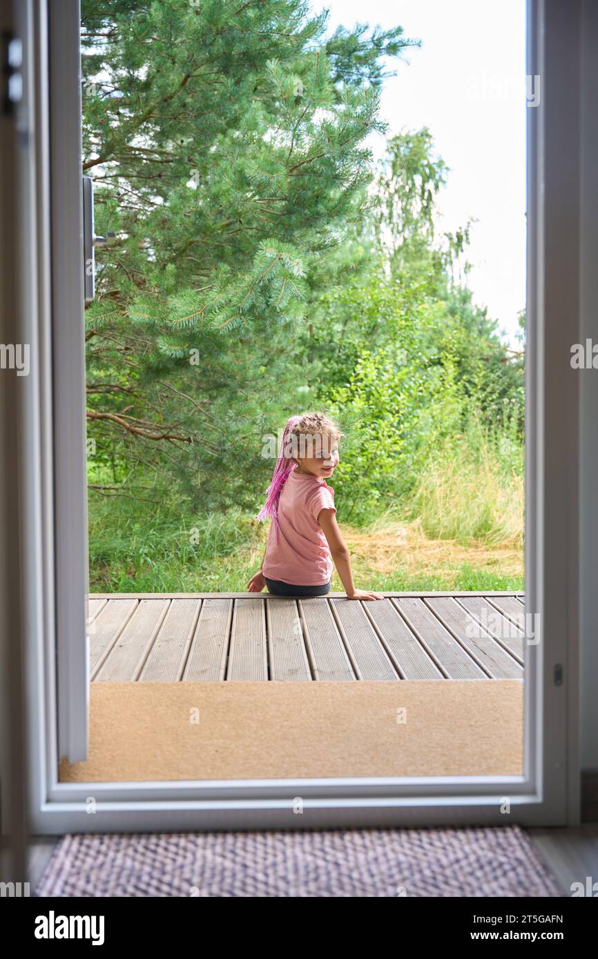 Bambina sorridente che si diverte a passatempo in una casa di campagna, seduto sul portico e guardando il paesaggio, respirando aria fresca nella foresta, trascorrendo le vacanze lontano Foto Stock