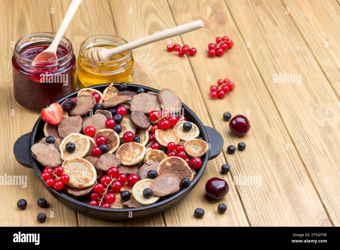 Pancake e frutti di bosco in padella. Miele e marmellata sul tavolo. Sfondo di legno chiaro. Vista dall'alto. Copia spazio Foto Stock