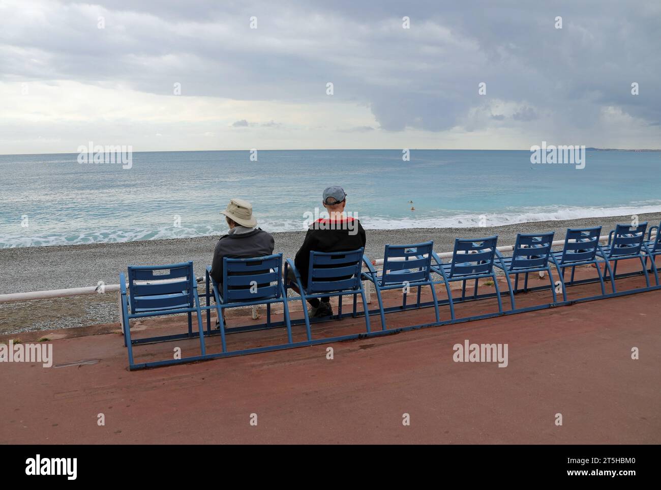 Turisti invernali seduti sulle famose sedie blu della Promenade des Anglais a Nizza Foto Stock