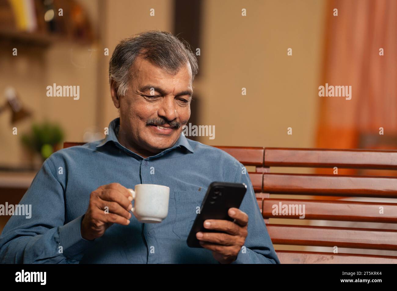 un uomo anziano sorridente sorridente che beve tè mentre usa il telefono cellulare a casa - concetto di relax mattutino, navigazione sui social media e tecnologia Foto Stock