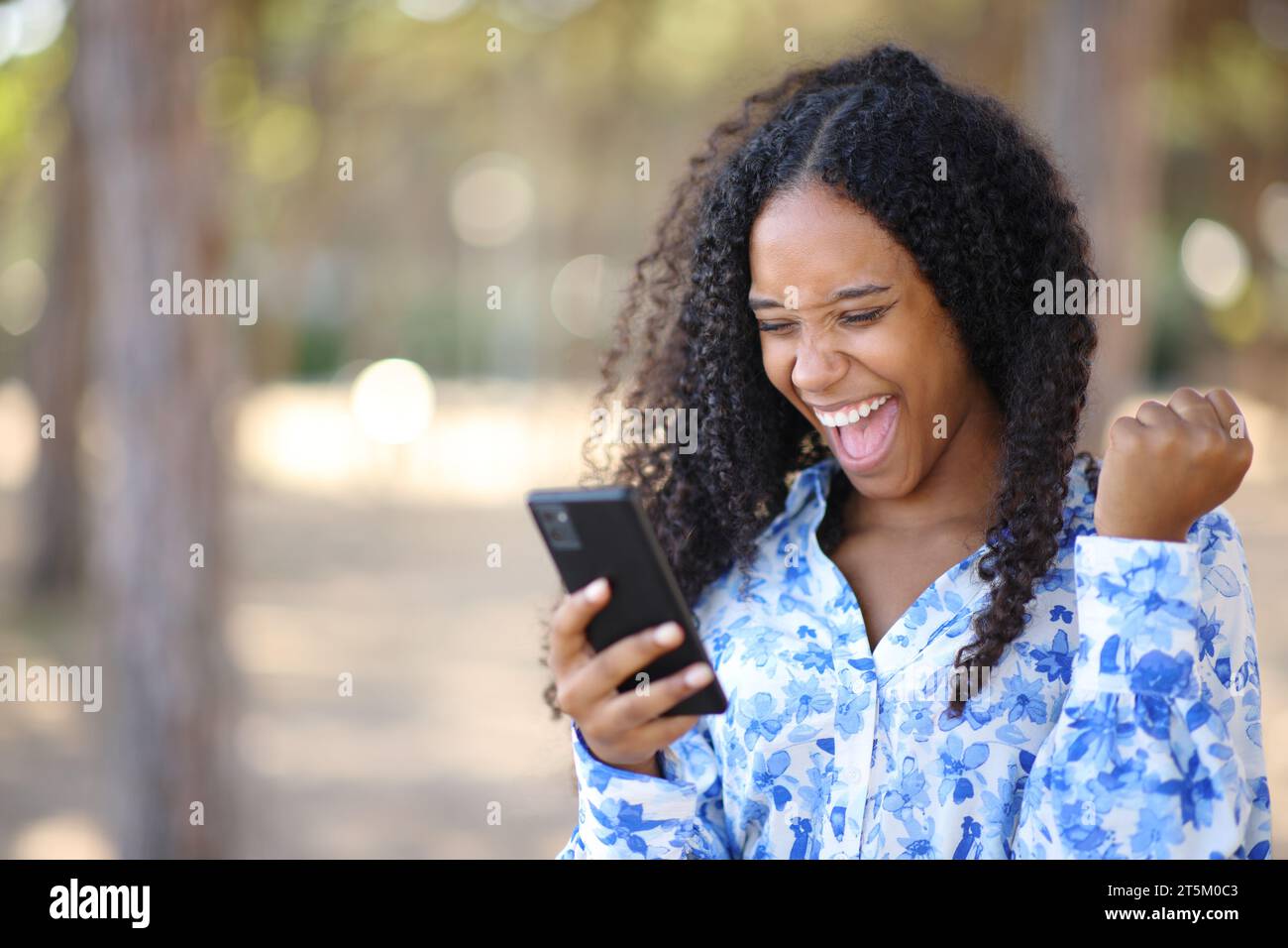 Eccitata donna nera che controlla il telefono e festeggia le buone notizie in un parco Foto Stock
