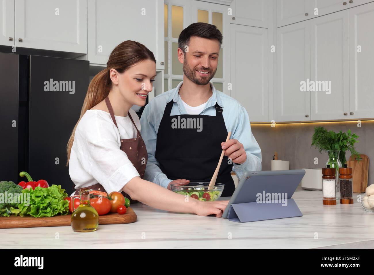Coppia felice che legge la ricetta su un tablet mentre cucina in cucina. Libro culinario online Foto Stock