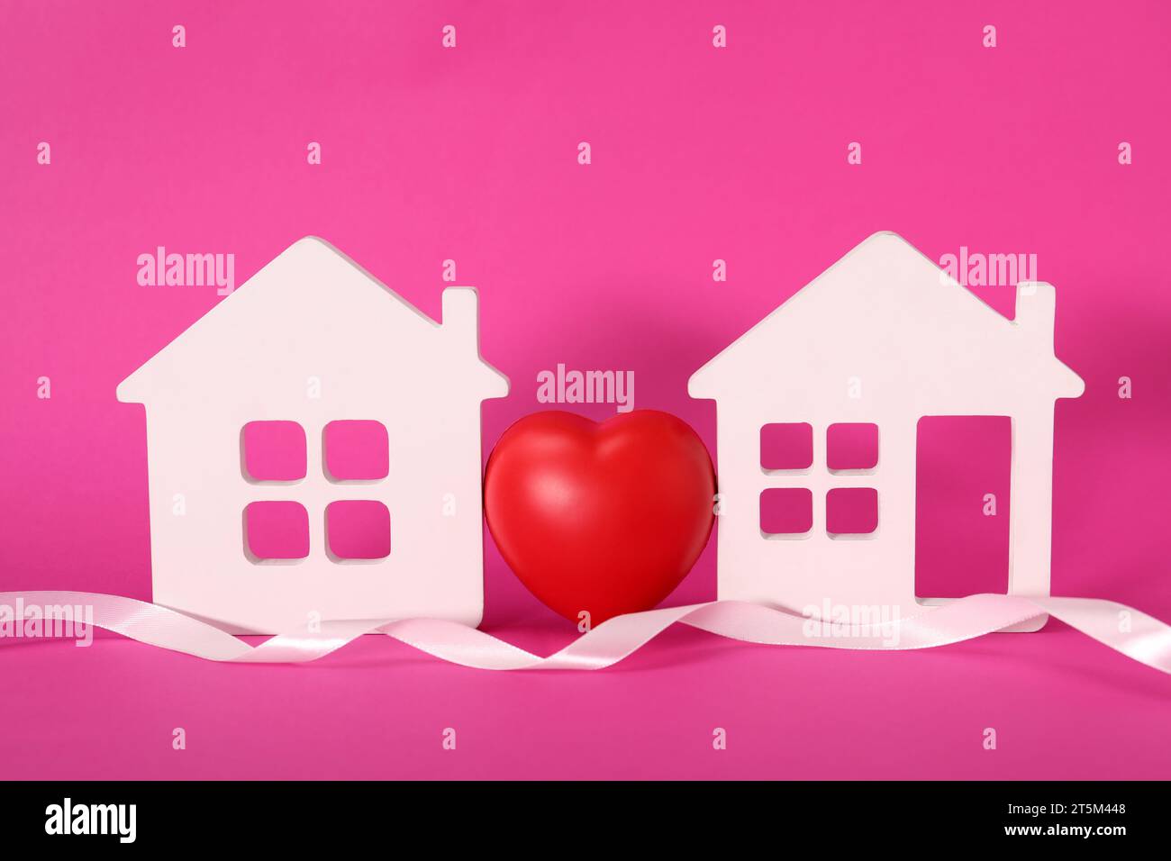 Nastro bianco e cuore decorativo tra due modelli di casa su sfondo rosa che simboleggia la connessione in relazione a lunga distanza Foto Stock