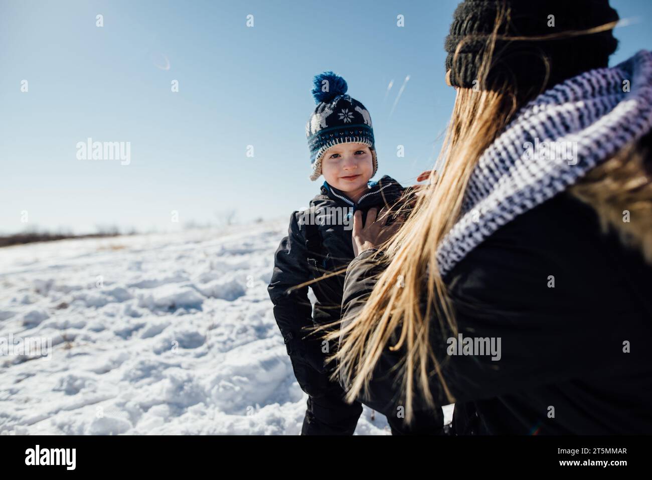 Vista posteriore della donna che aiuta il bambino con i pantaloni da neve mentre è piccolo Foto Stock