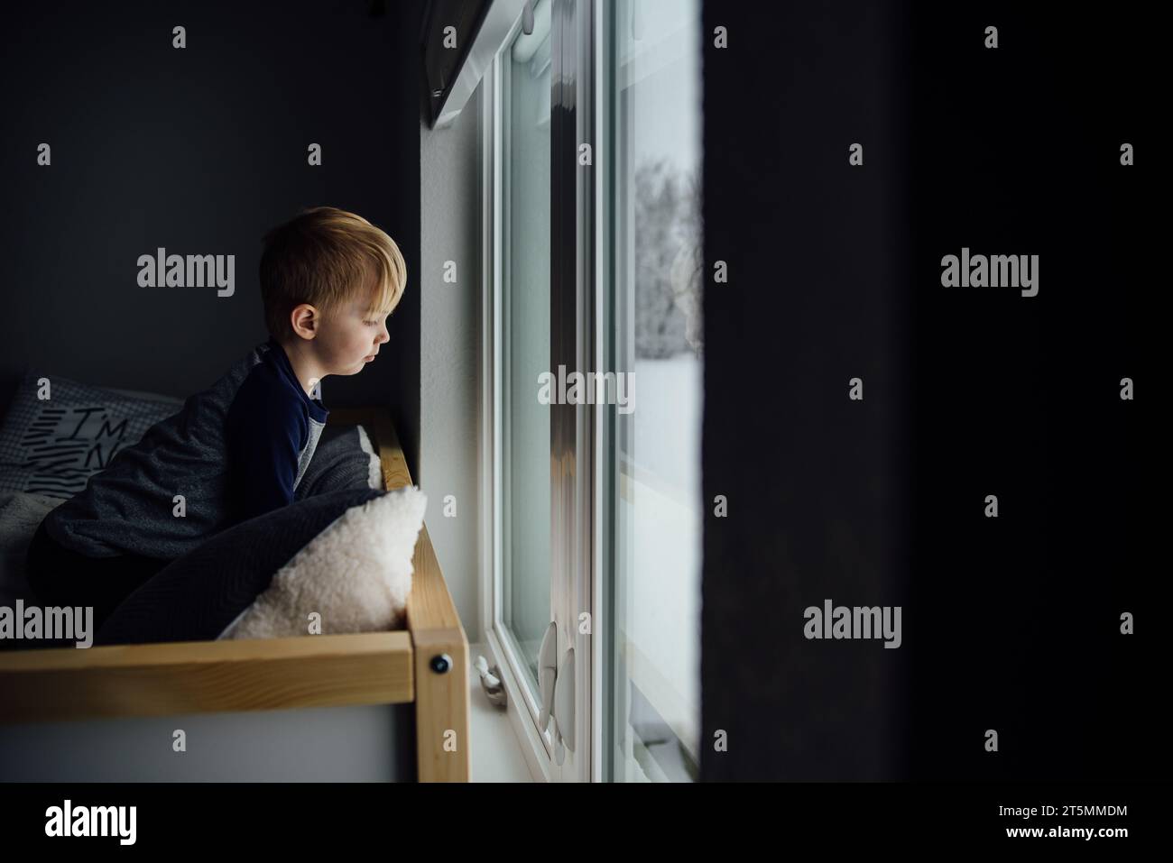 Un bambino seduto sul letto e guarda fuori dalla finestra sulla neve sotto con dr Foto Stock