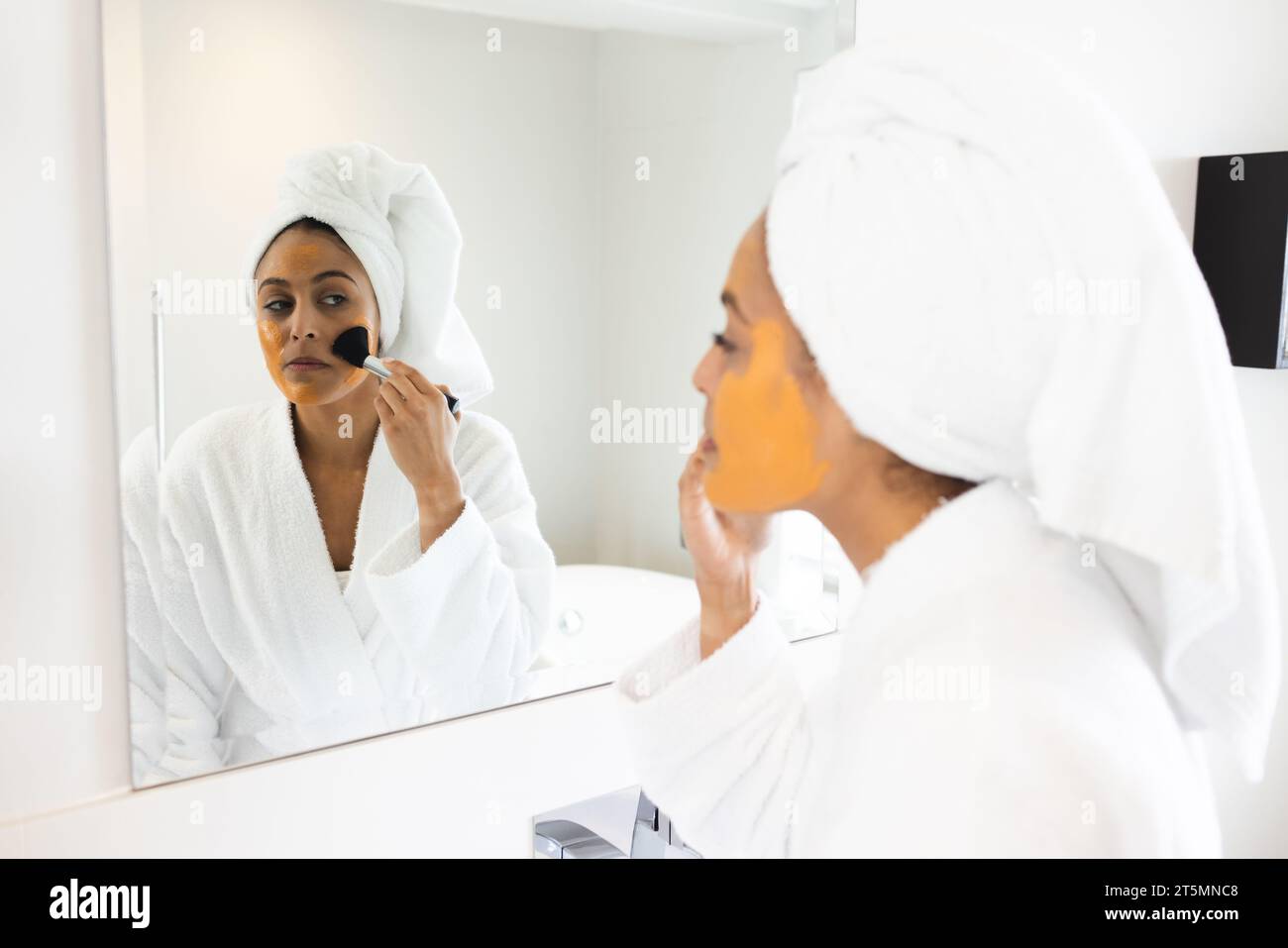 Felice donna birazziale che indossa accappatoio e fa una maschera di fronte allo specchio in bagno Foto Stock