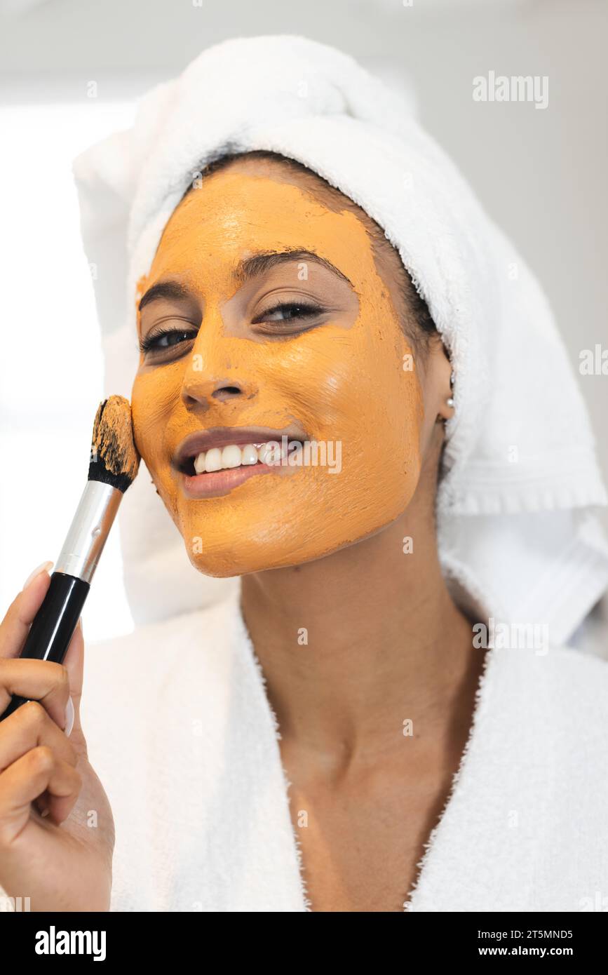 Ritratto di felice donna birazziale che indossa accappatoio bianco e fa la maschera facciale in bagno Foto Stock