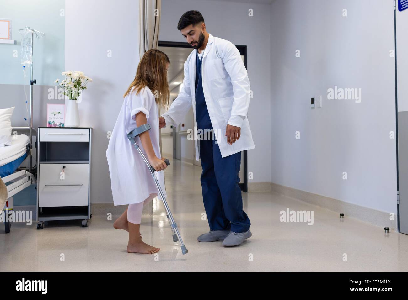 Medico maschile diversificato osservando la ragazza uso del paziente stampelle in reparto ospedaliero, copia spazio Foto Stock