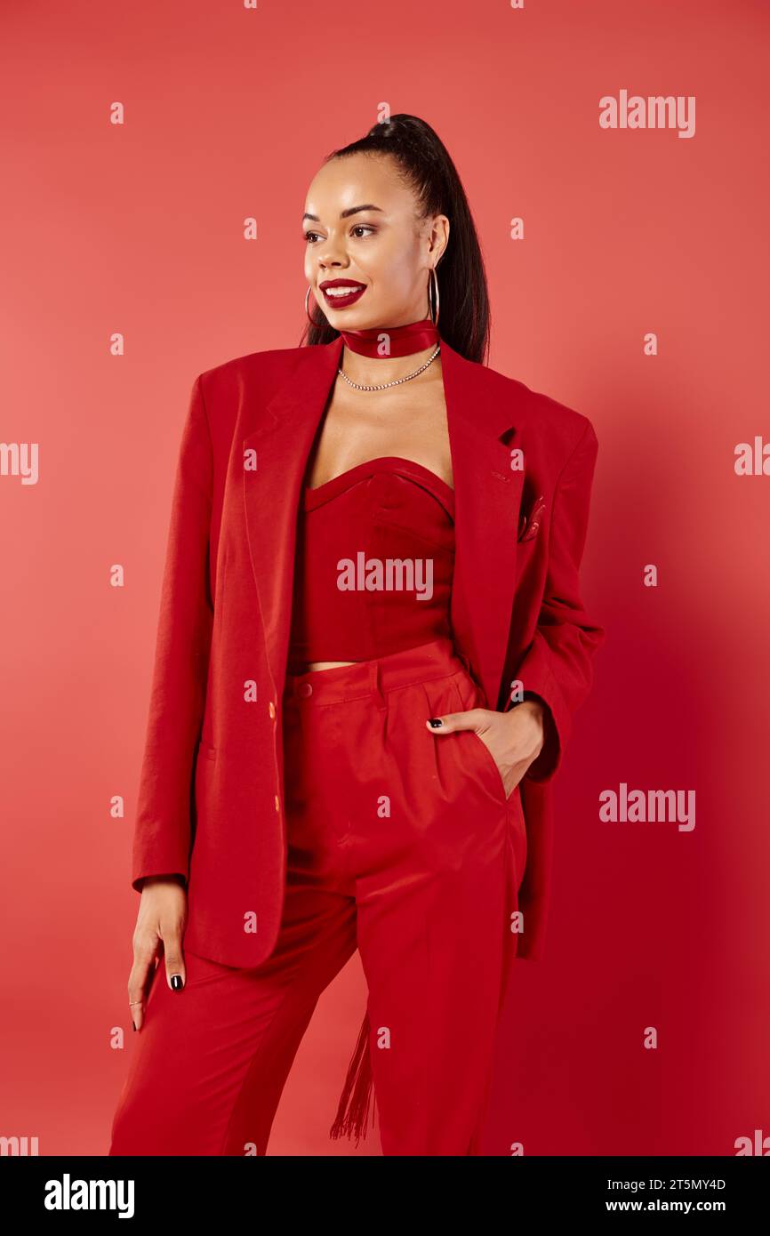 felice donna afro-americana con coda di cavallo in posa con la mano in tasca di pantaloni su sfondo rosso Foto Stock