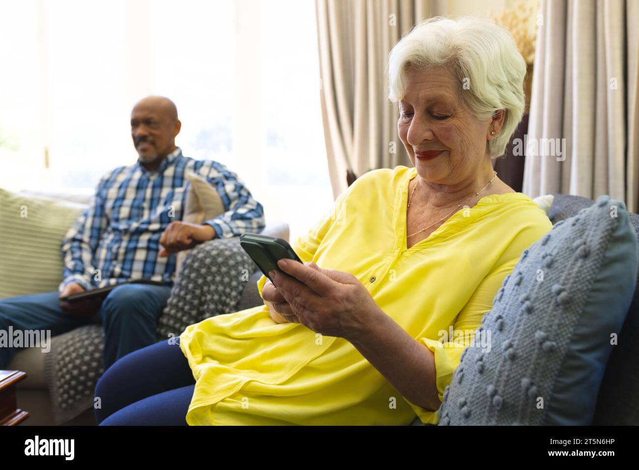 Donna anziana caucasica felice seduta sul divano, con smartphone e sorridendo nel soleggiato soggiorno Foto Stock