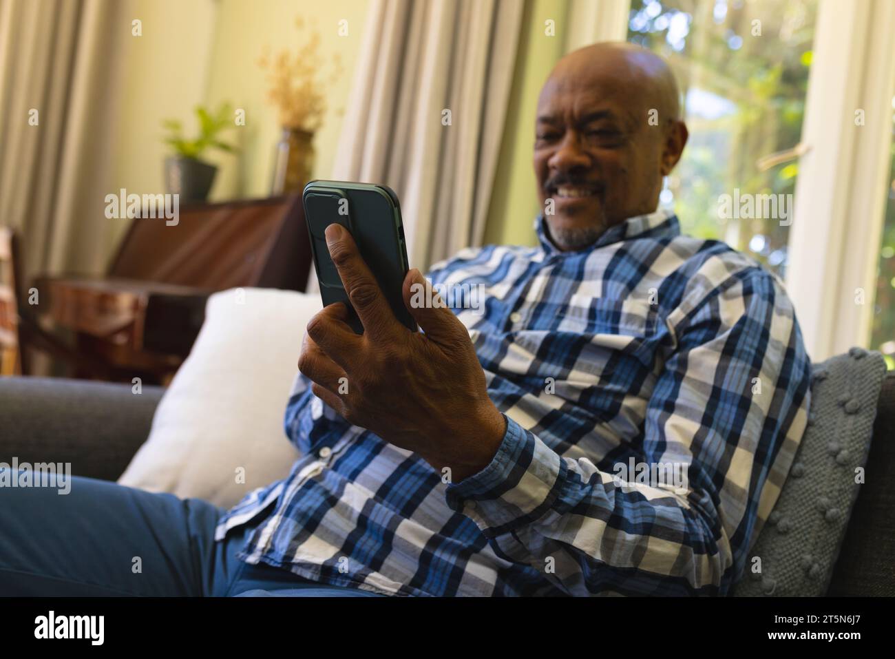 Felice anziano afro-americano seduto sul divano, usando lo smartphone e sorridendo nel soleggiato soggiorno Foto Stock