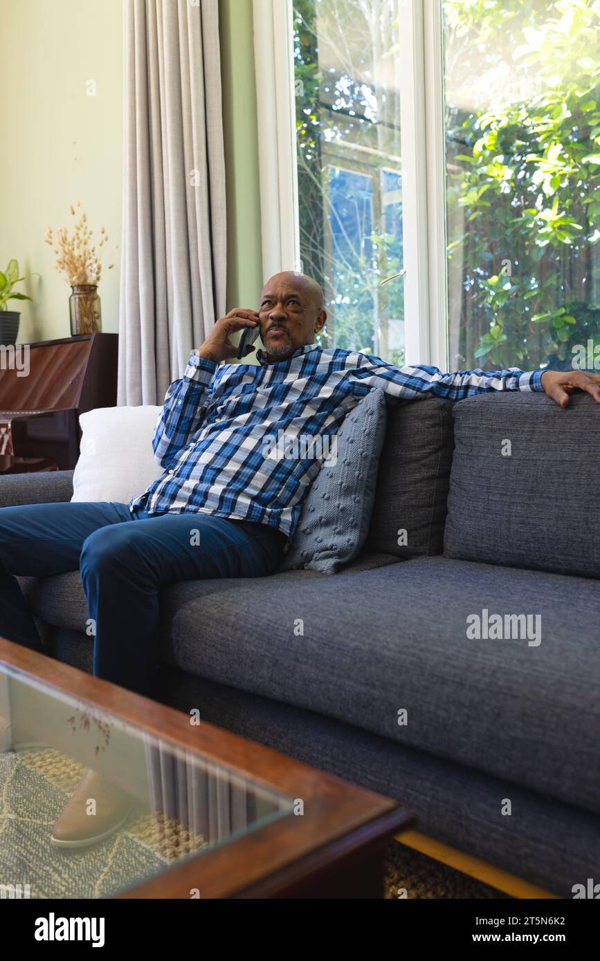 Felice anziano afro-americano seduto sul divano e utilizzando lo smartphone nel soleggiato soggiorno Foto Stock