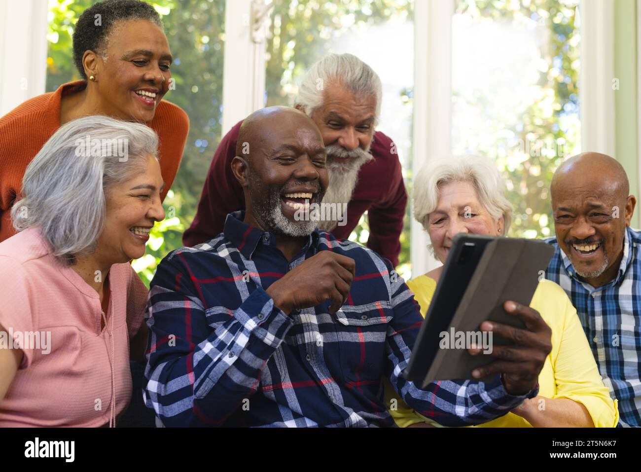 Felice e diversificato gruppo di amici anziani che videochiama e ride nel soleggiato salotto Foto Stock