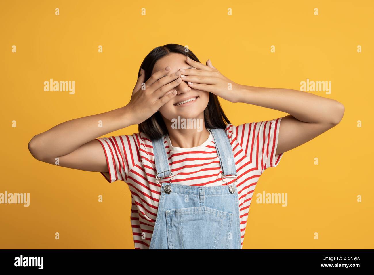 Ragazza adolescente felice che chiude gli occhi con le mani in posa su sfondo giallo giocando a nascondino. Foto Stock