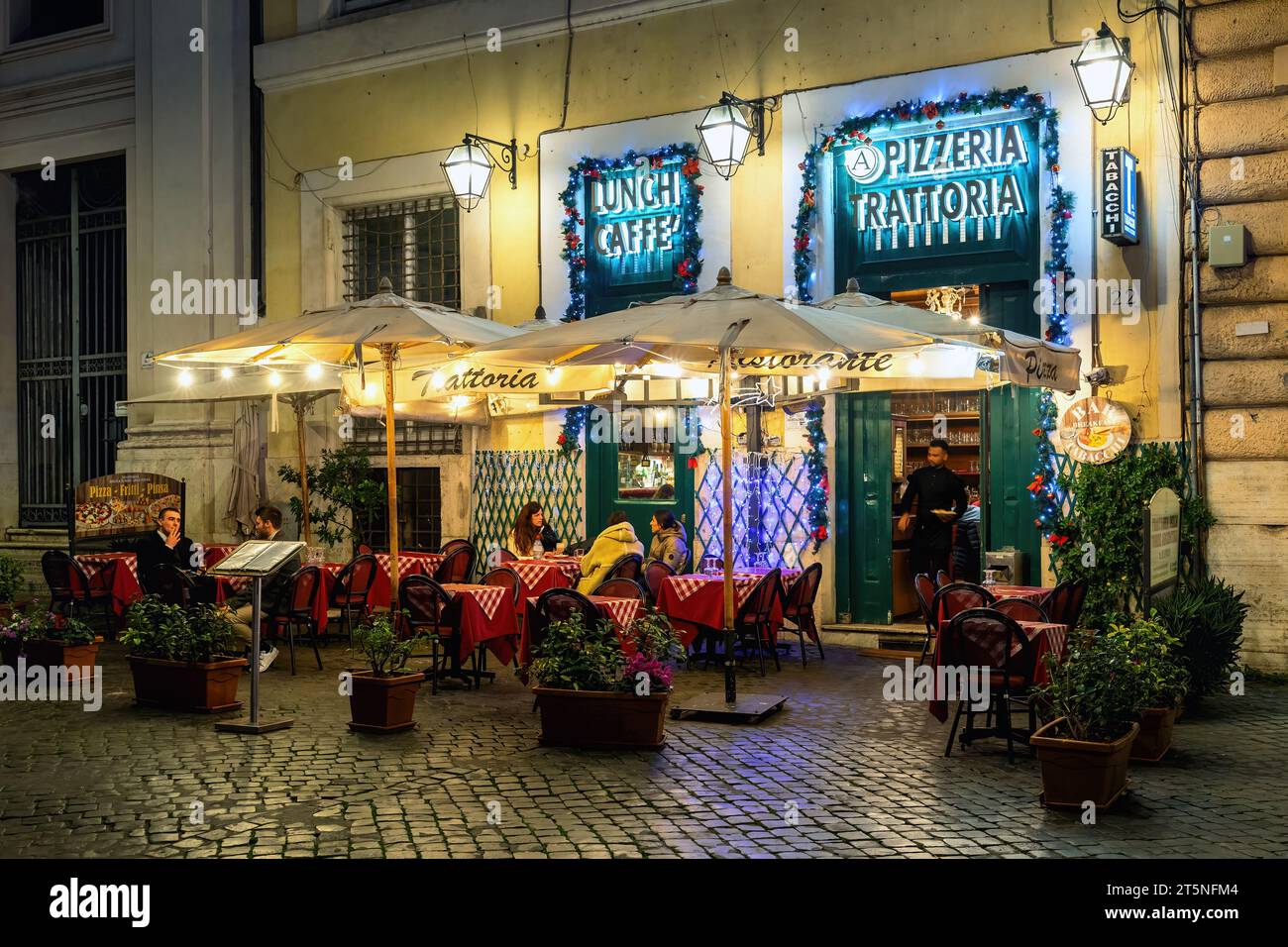 Persone sedute in un piccolo ristorante all'aperto su una strada acciottolata illuminata la sera a Roma, Italia. Foto Stock