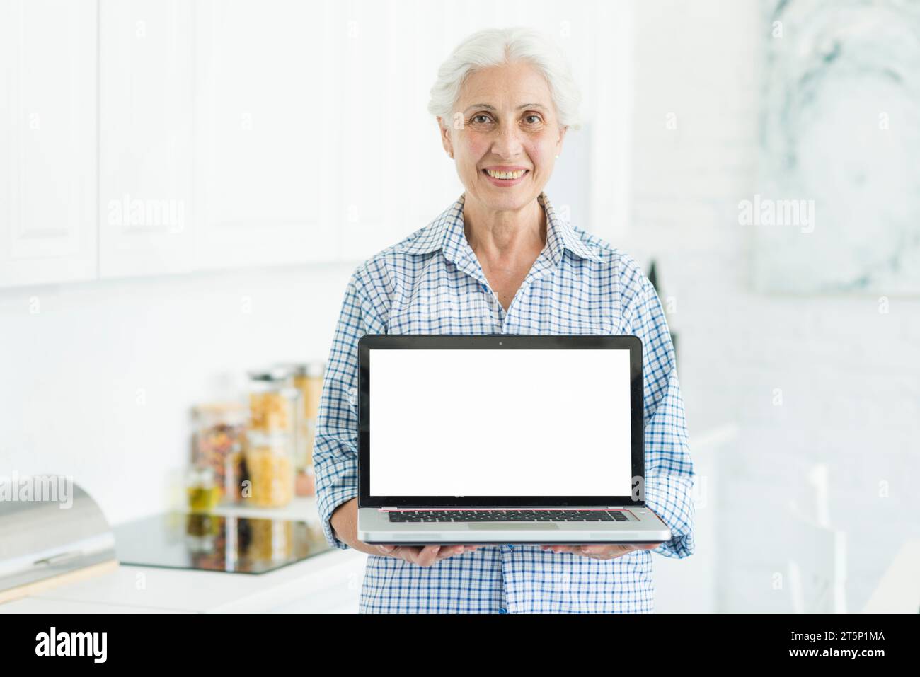 Donna anziana sorridente in cucina che mostra un notebook con schermo bianco Foto Stock