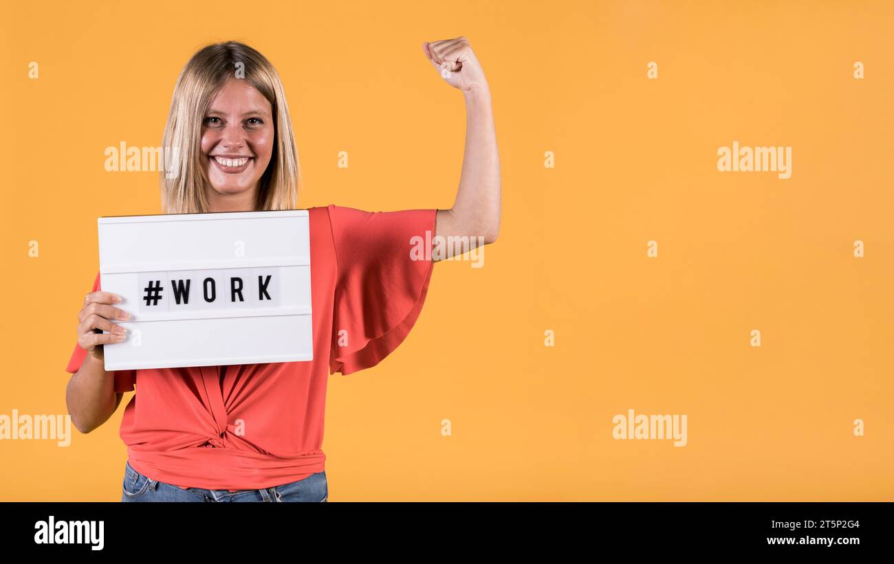 Giovane donna sorda che tiene una scatola leggera con hash tag lavoro testo flessibile muscolare sfondo giallo Foto Stock