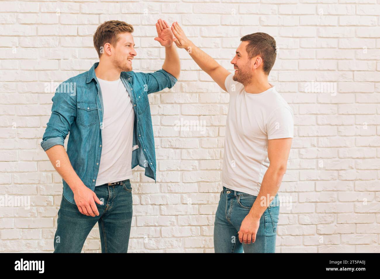 Due amici maschi che si danno cinque in alto l'uno contro il muro di mattoni bianchi Foto Stock