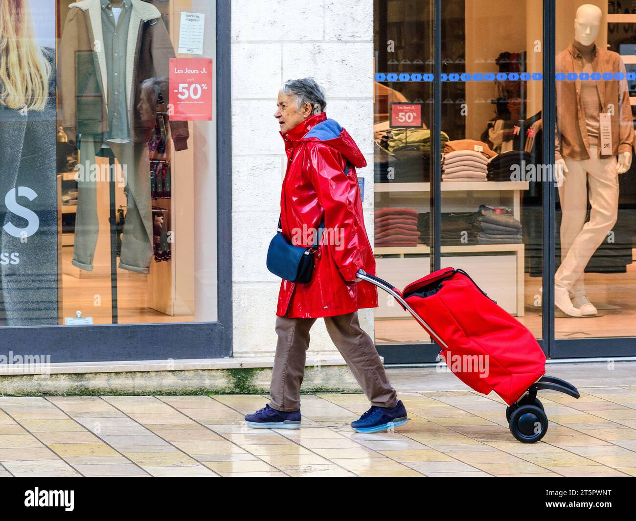 Donna anziana in plastica rossa impermeabile che tira il carrello rosso della spesa lungo la pavimentazione della città - Tours, Indre-et-Loire (37), Francia. Foto Stock
