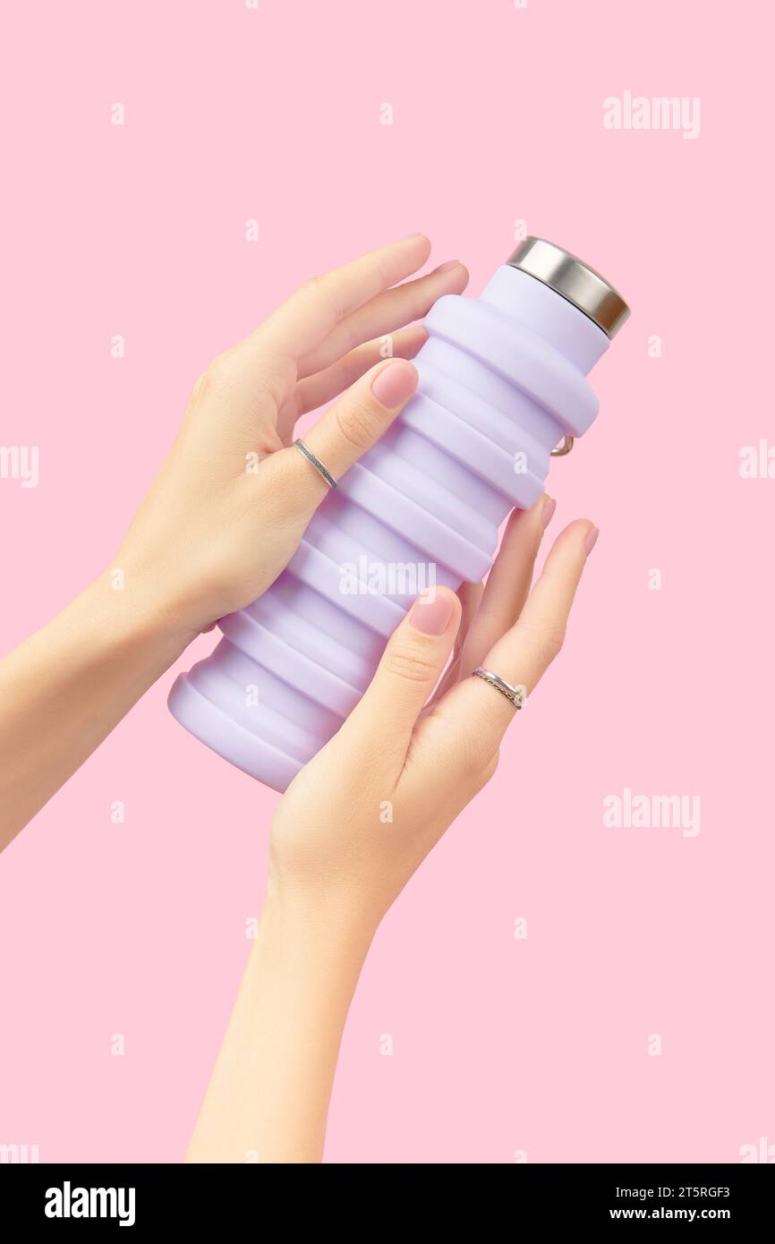Mani femminili che reggono una bottiglia d'acqua lilla riutilizzabile pieghevole su sfondo rosa. Stile di vita sostenibile Foto Stock