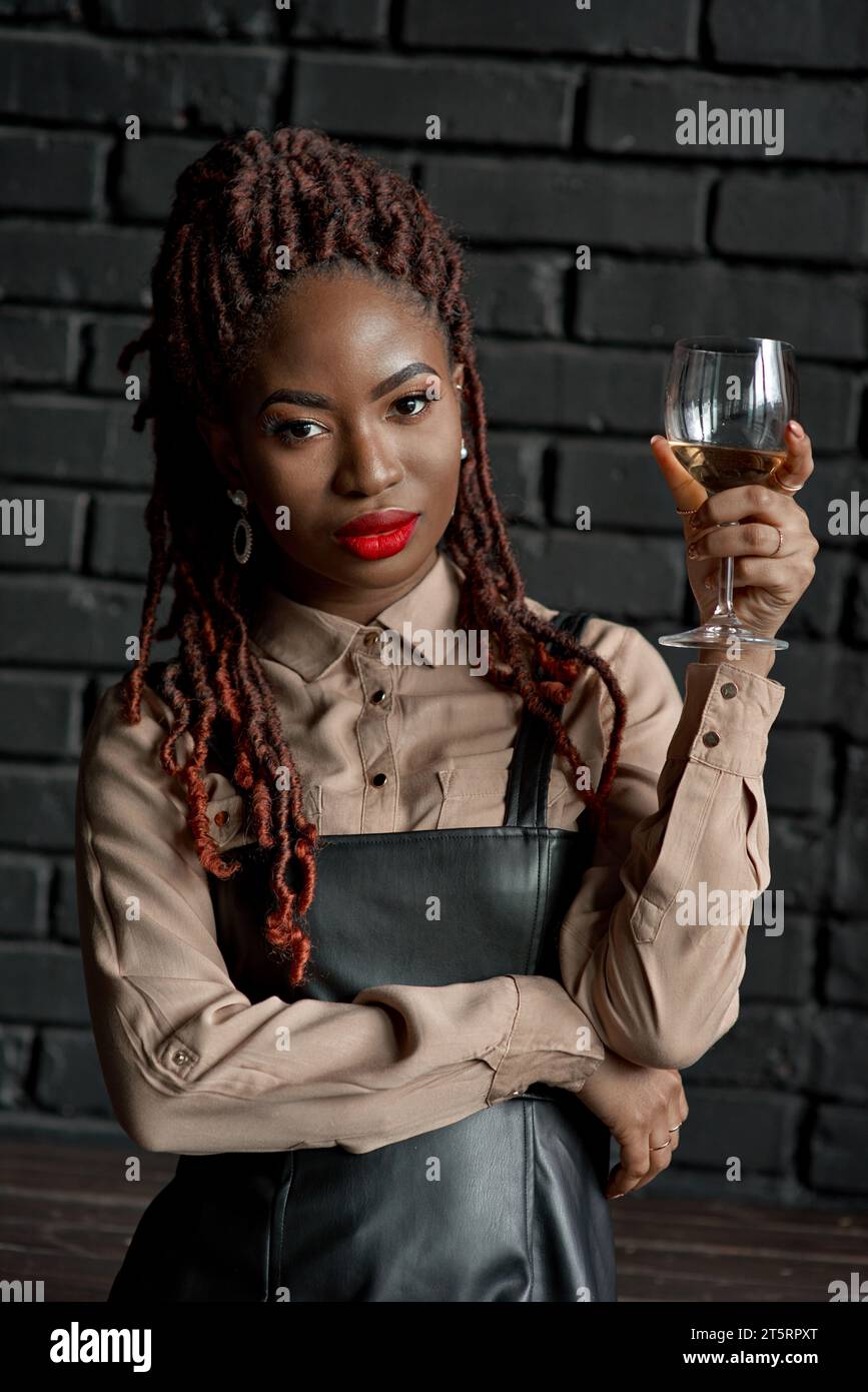 Un ritratto ravvicinato a mezzo corpo di una donna africana sicura di sé e ben vestita che regge un bicchiere di vino bianco su uno sfondo di mattoni neri. Degustazione di vini, Foto Stock
