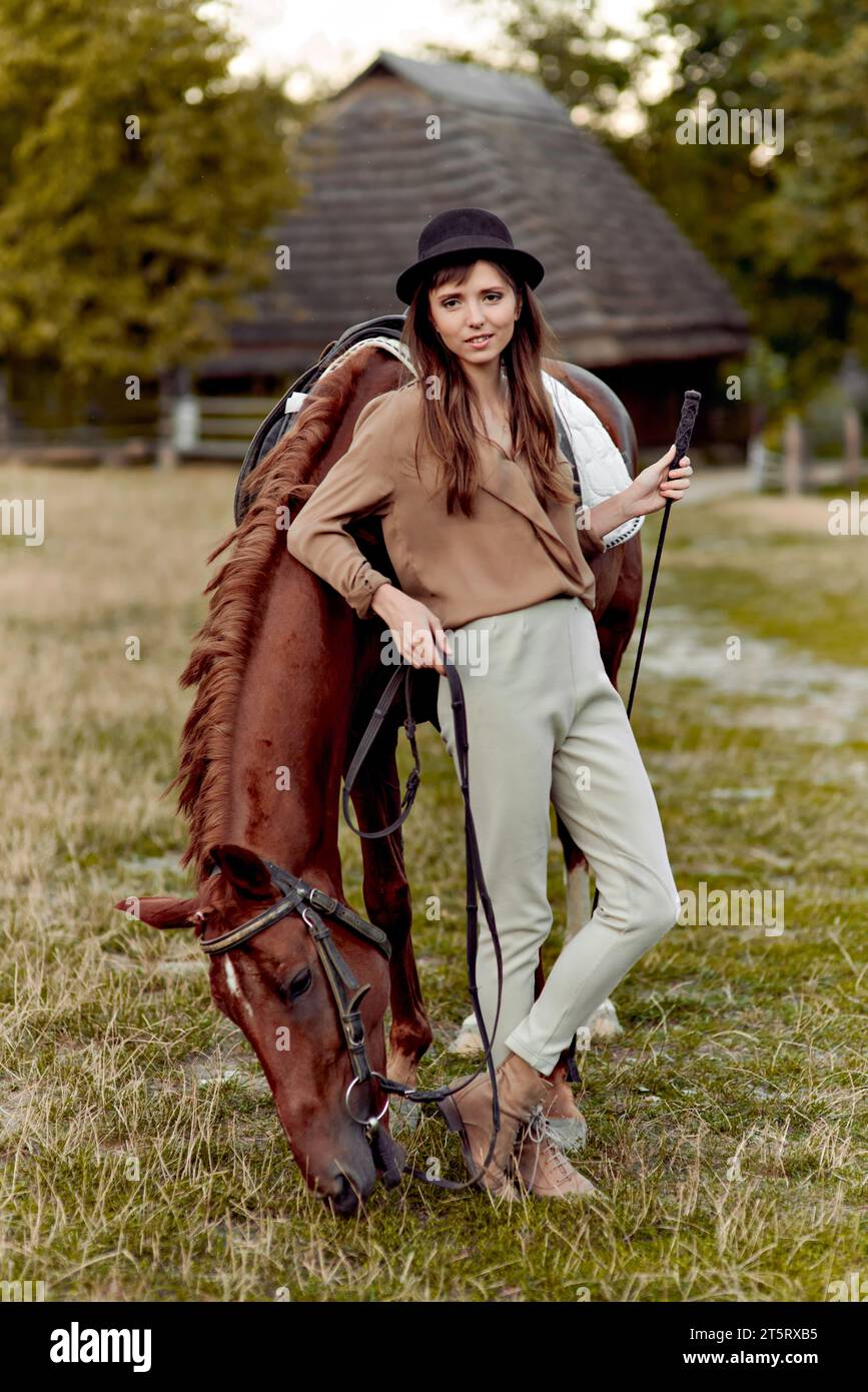 Giovane donna con cappello nero si trova sull'erba vicino a un cavallo bruno che pascolava l'erba. Cavalli che vengono nutriti. Buono omaggio per ippoterapia ed emozioni. Cavallo dentro Foto Stock