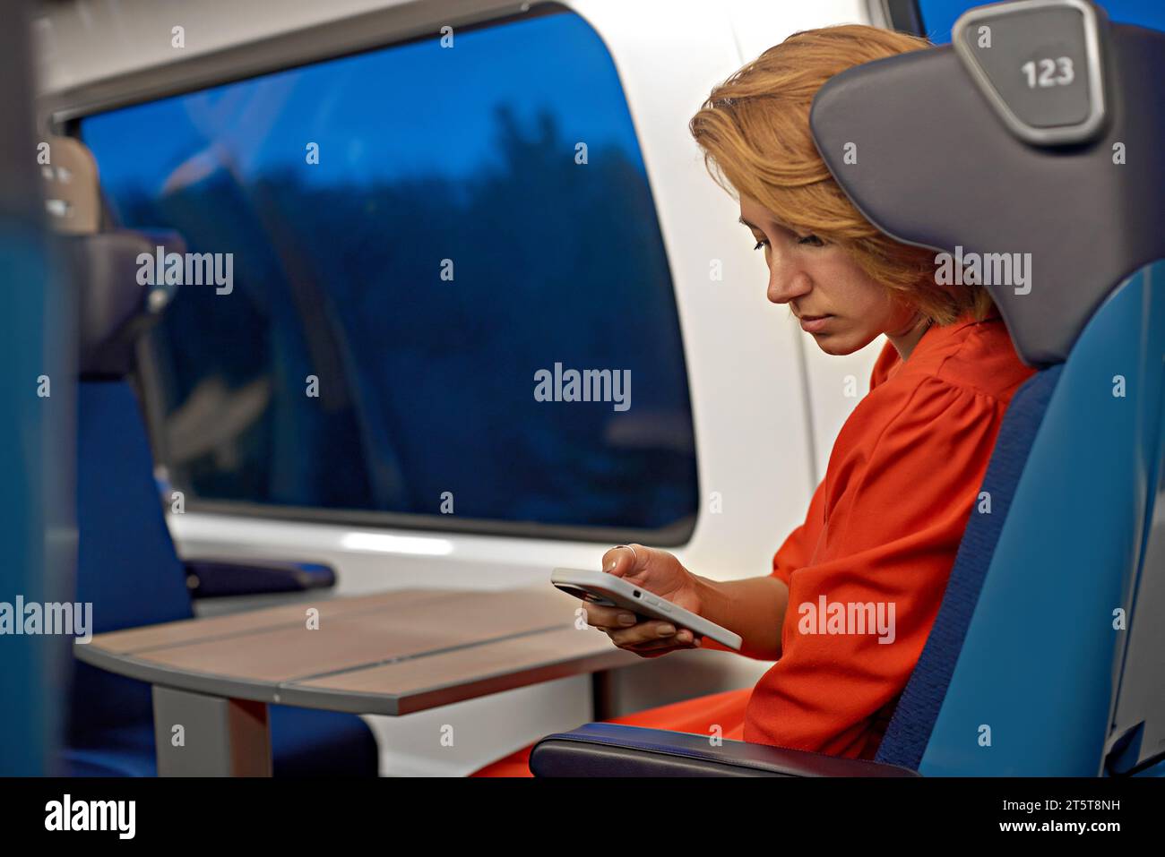 Una giovane donna attraente con i capelli rossi e un vestito rosso che lavora online in remoto dal telefono e viaggia. Viaggi in treno e lettura sul telefono cellulare Foto Stock