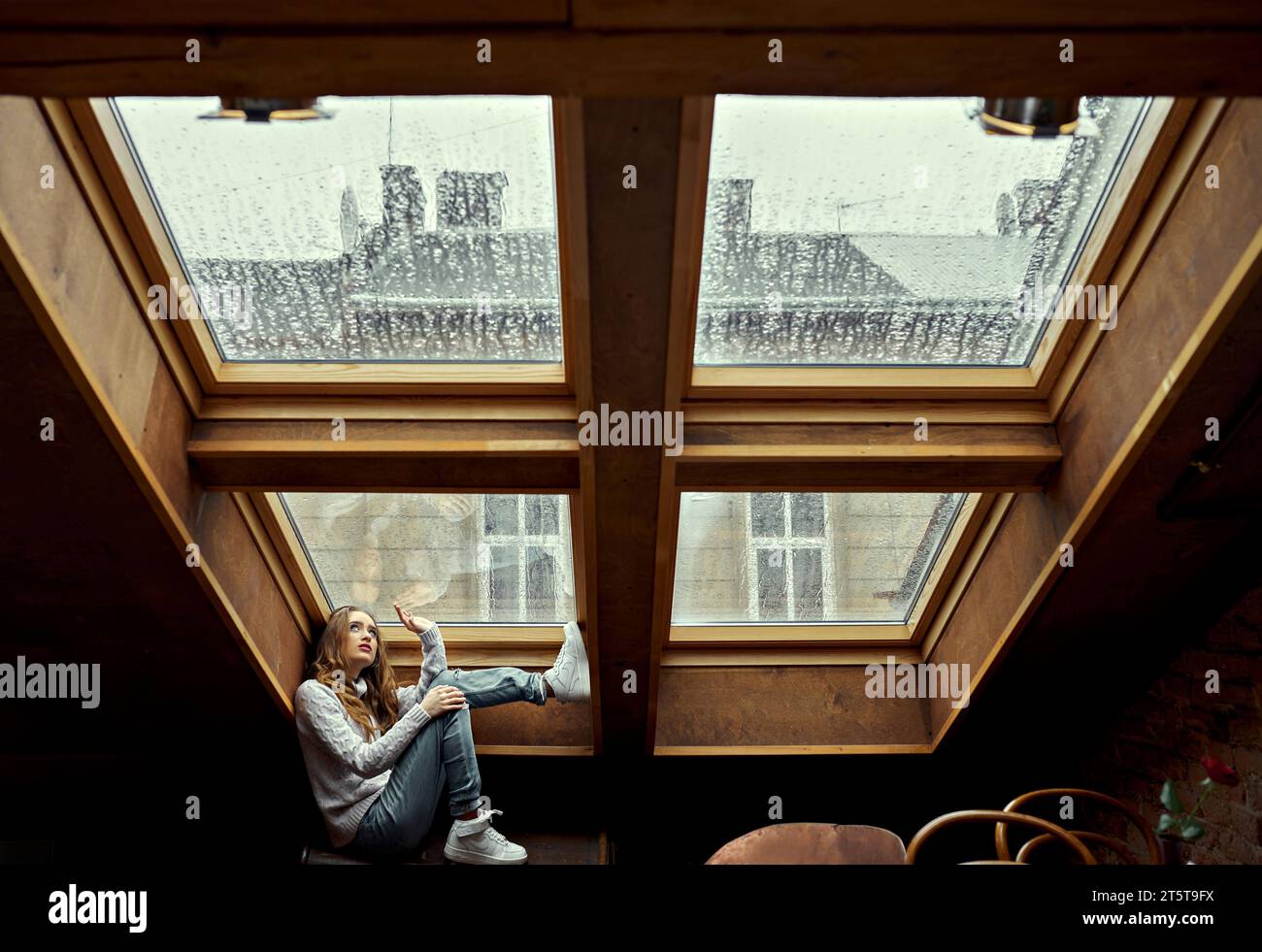Una ragazza con un maglione bianco guarda in alto, toccando la finestra nel soffitto della casa con una mano. Autunno e pioggia fuori dalla finestra. Il concetto di Foto Stock