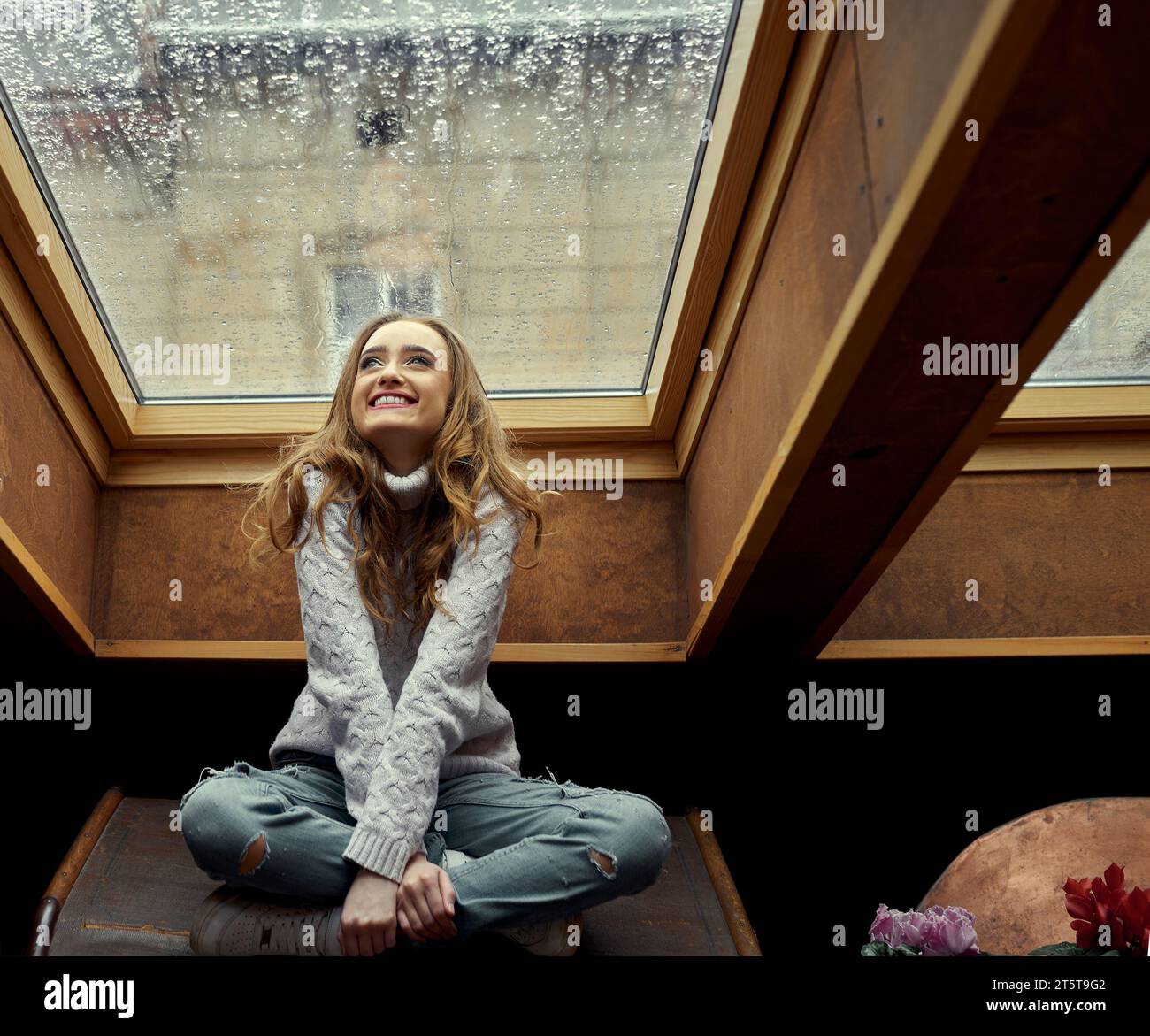 La giovane ragazza è felice della pioggia fuori dalla finestra, seduta nella sua accogliente stanza sotto la finestra nel soffitto, su cui scorre la pioggia. Concetto di io Foto Stock