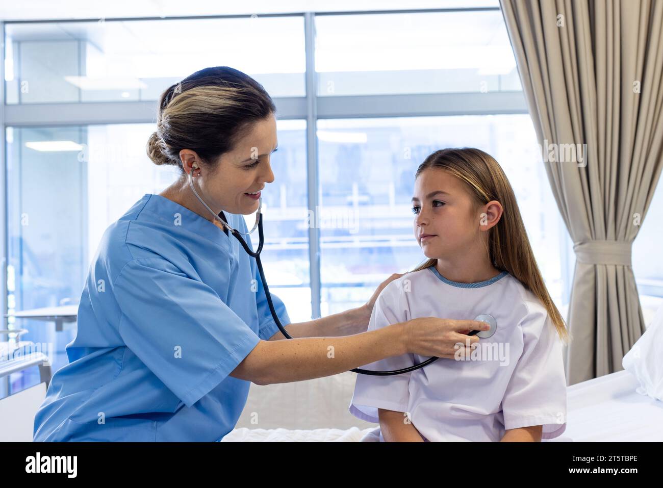 Medico femminile caucasico sorridente che utilizza stetoscopio sul torace di paziente ragazza seduto sul letto d'ospedale Foto Stock