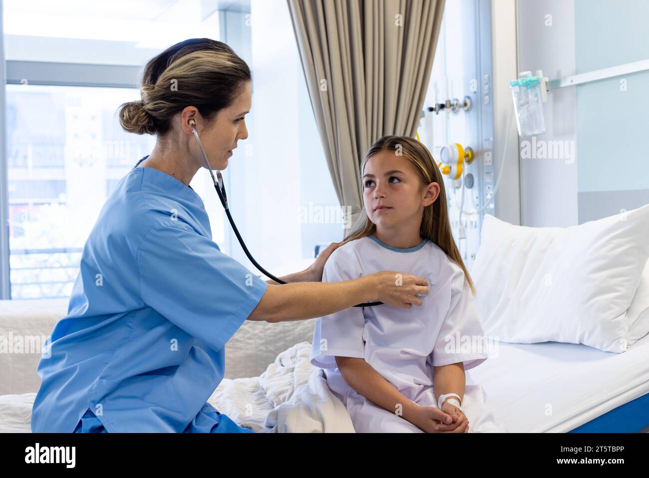 Medico femminile caucasico focalizzato che utilizza stetoscopio sul torace della ragazza paziente seduta sul letto d'ospedale Foto Stock