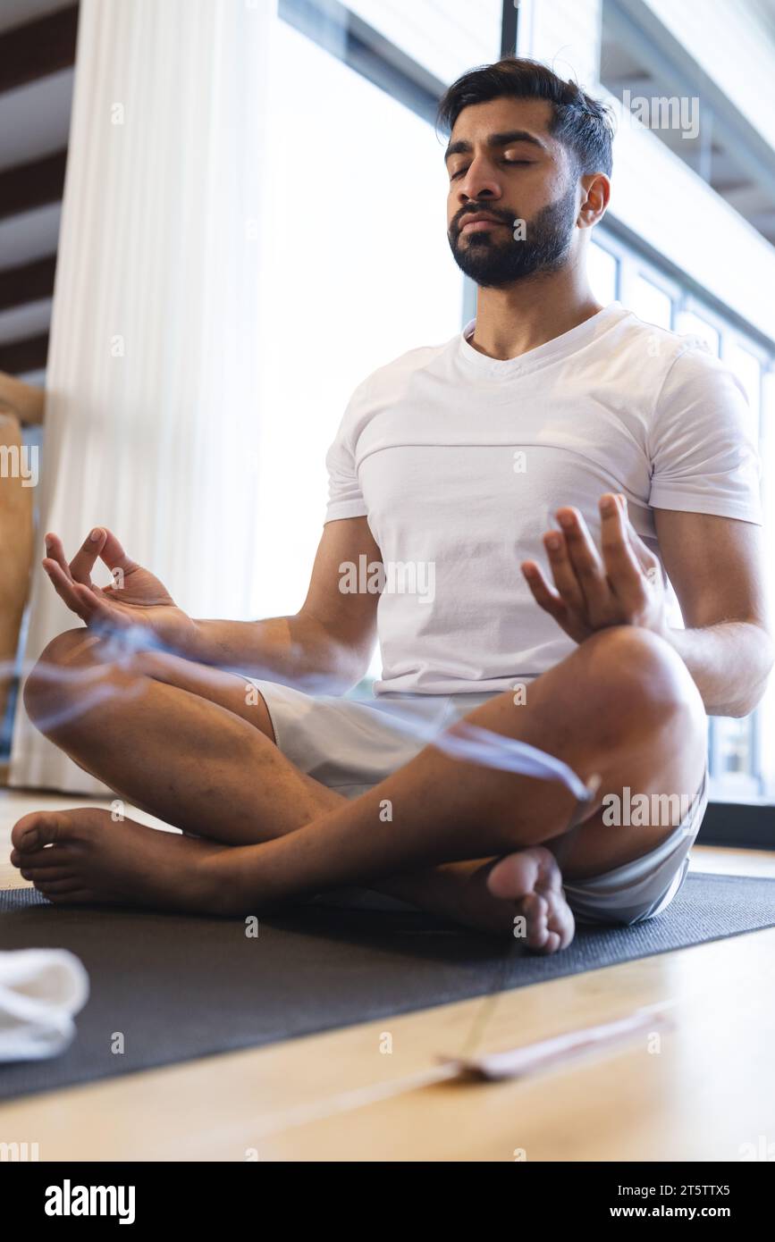 Rilassato uomo birazziale seduto, praticando yoga, chiudendo gli occhi e meditando in una stanza soleggiata a casa Foto Stock