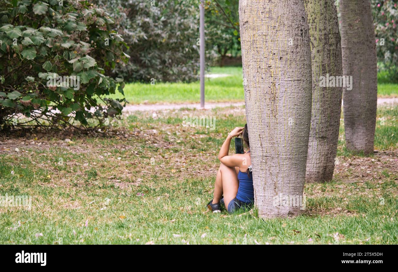 Una giovane donna seduta contro un albero in un parco pubblico che guarda il suo cellulare Foto Stock