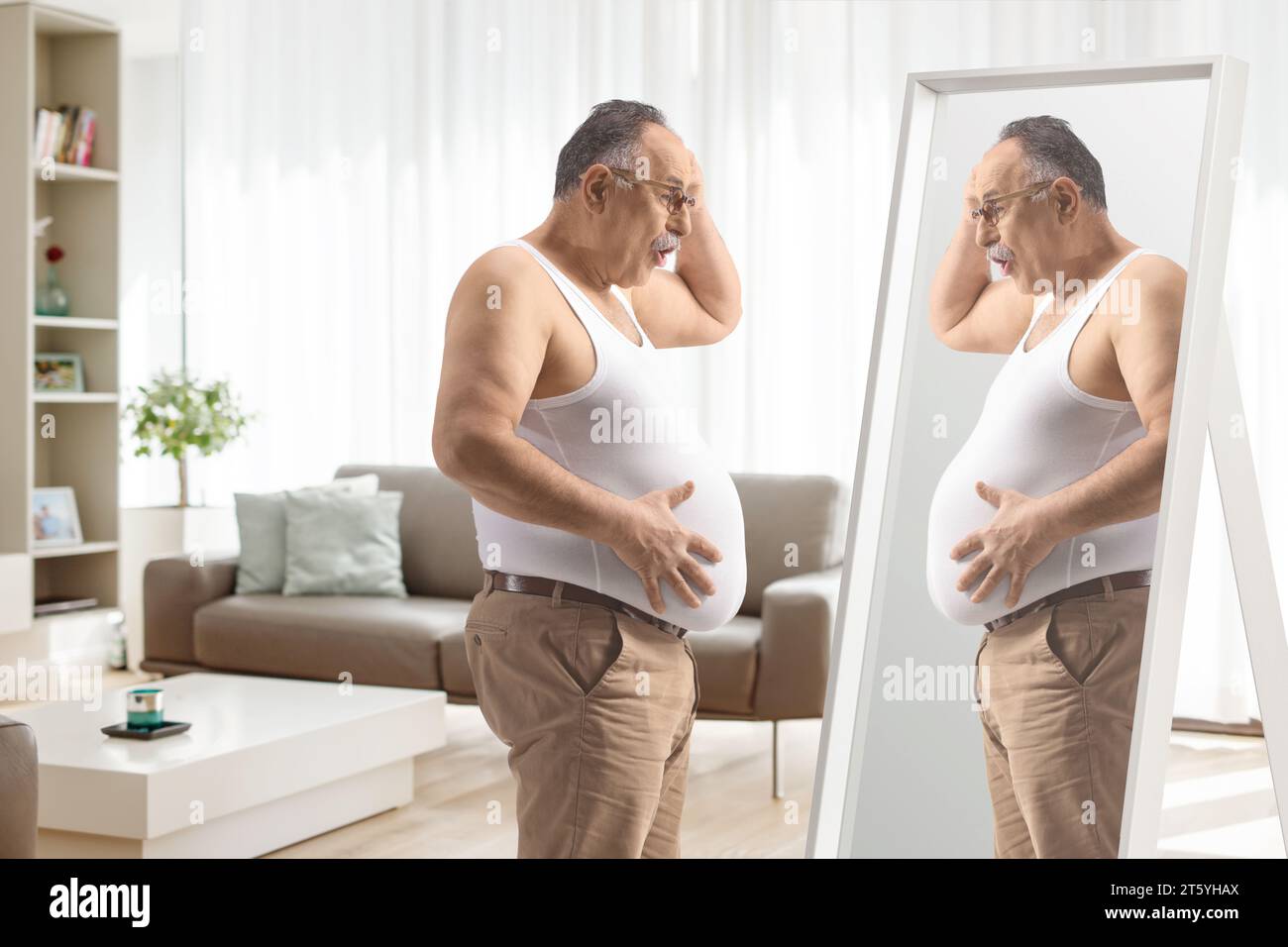 Un uomo maturo scioccato che guarda la sua grande pancia nello specchio all'interno di un salotto Foto Stock