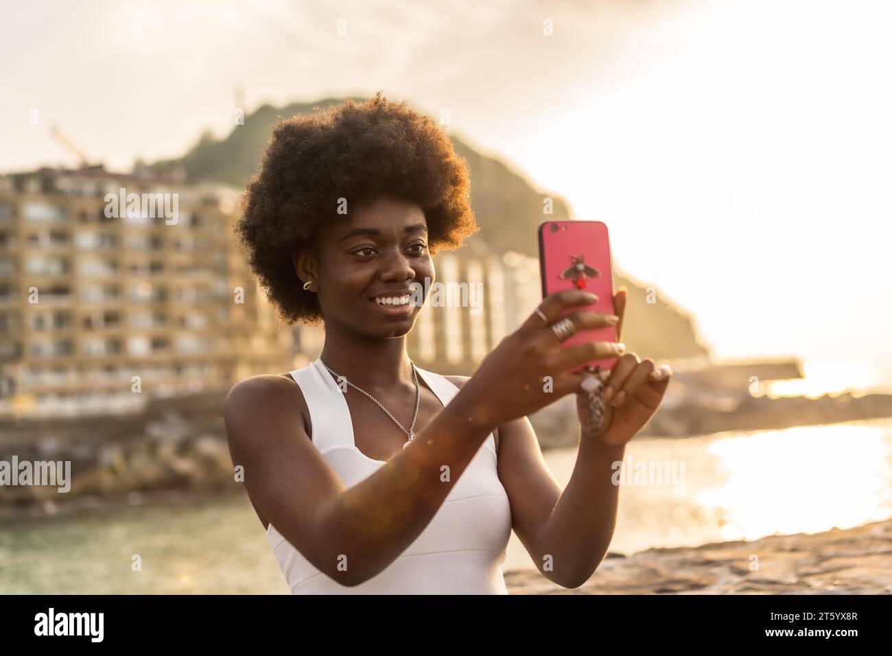 Felice donna afro in abito bianco che scatta un selfie al tramonto vicino al mare Foto Stock
