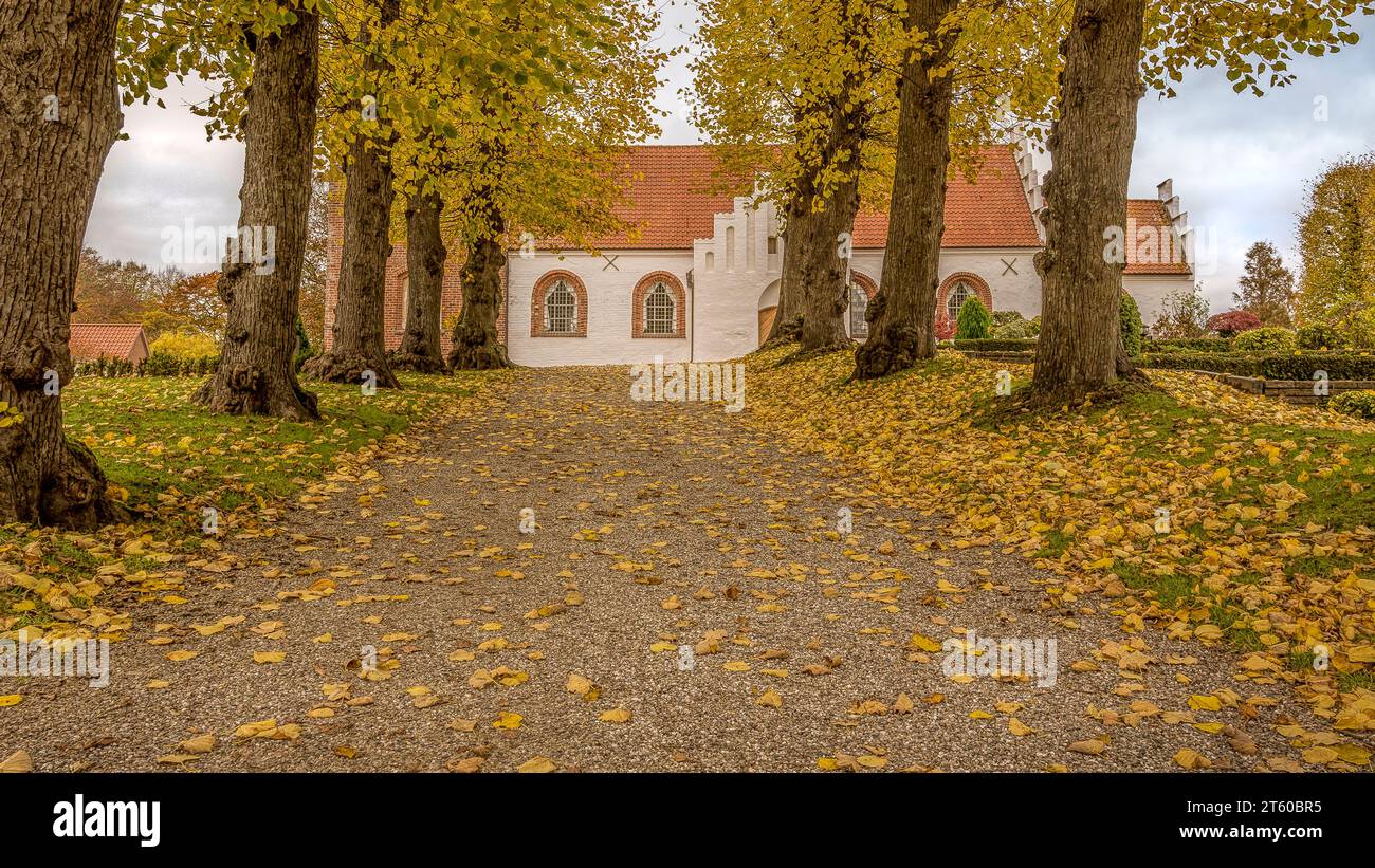 Un lungo sentiero di ghiaia con foglie cadute che conduce ad una chiesa bianca a Helsinge, Danimarca, il 4 novembre 2023 Foto Stock