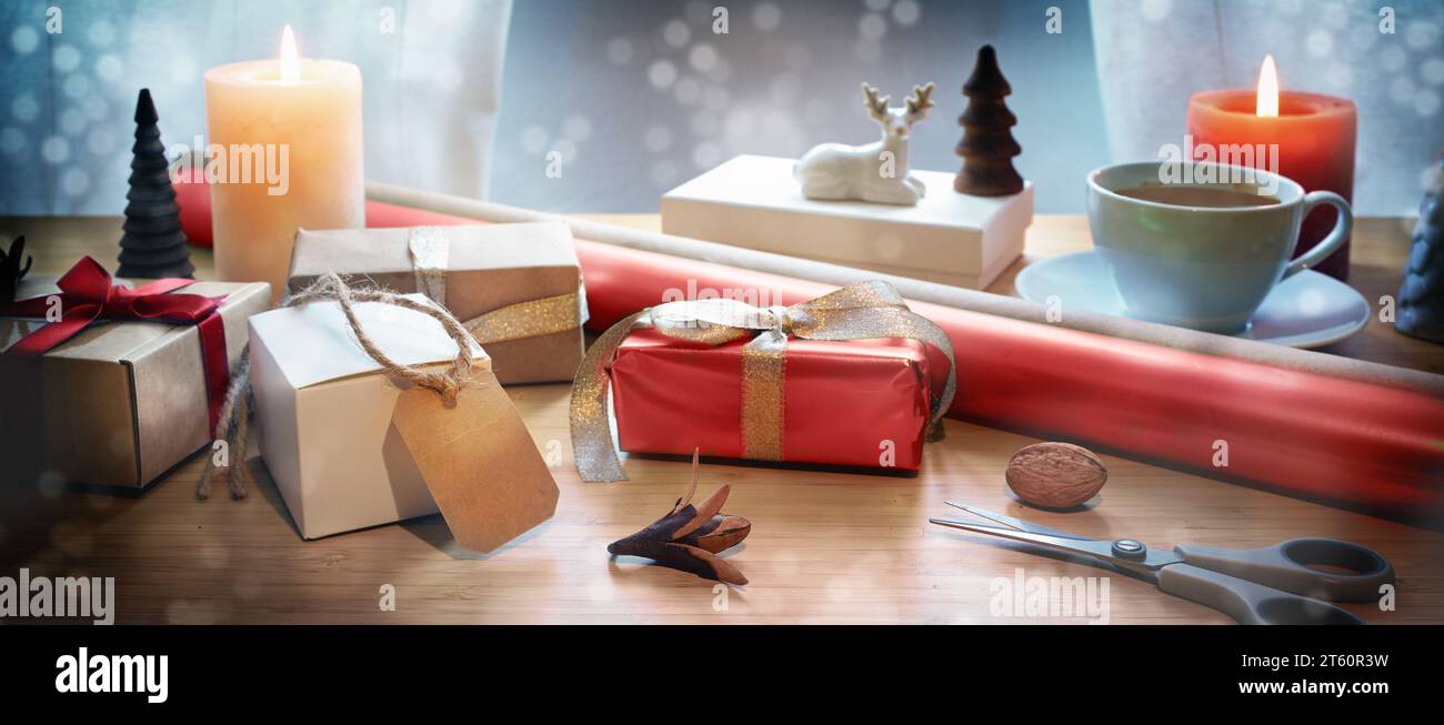 Confezione regalo natalizia su un tavolo con scatole regalo, forbici, carta rossa, candele e decorazioni natalizie, bokeh innevato, formato panoramico, spazio fotocopie, Foto Stock