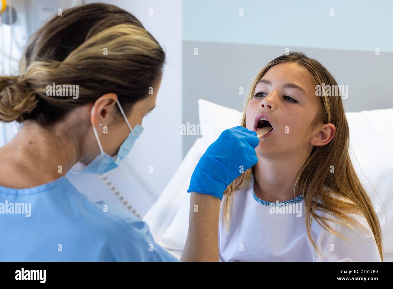 Medico femminile caucasico in maschera e guanti utilizzando spatola controllo gola di ragazza paziente in ospedale Foto Stock